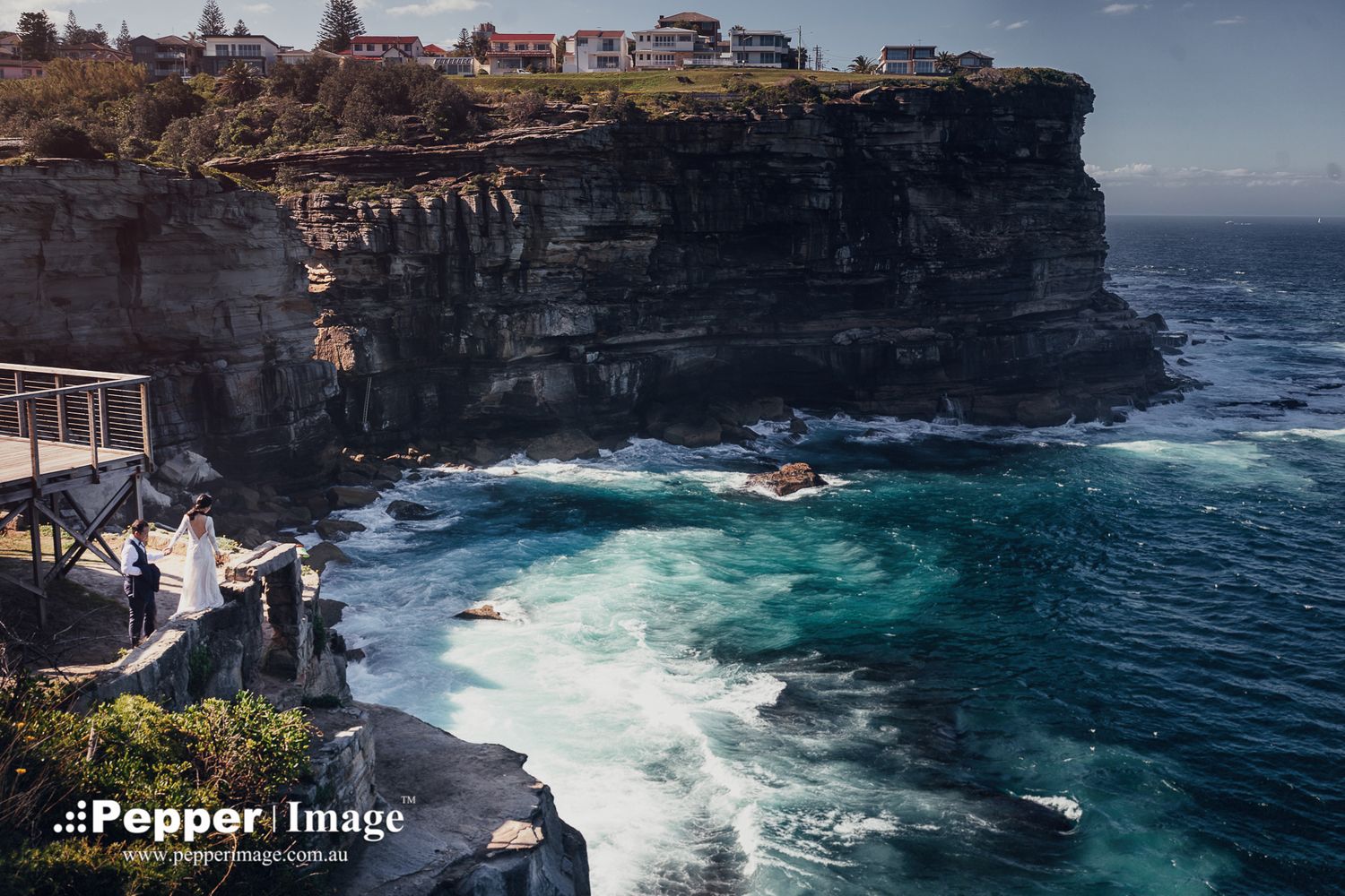 Aerial view of bride and groom on rocky coastal cliff with crashing ocean waves.