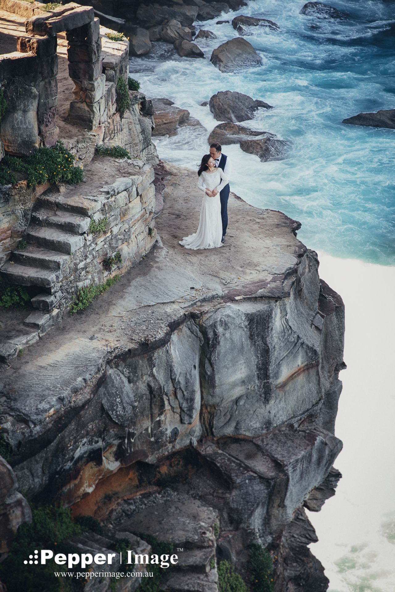 Bride and groom standing on dramatic coastal cliff edge with turquoise ocean waves below.