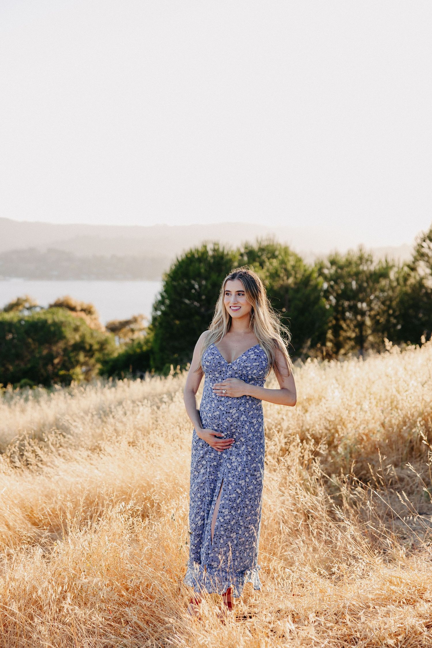 Pregnant woman in blue floral dress standing in golden grass with water view behind.