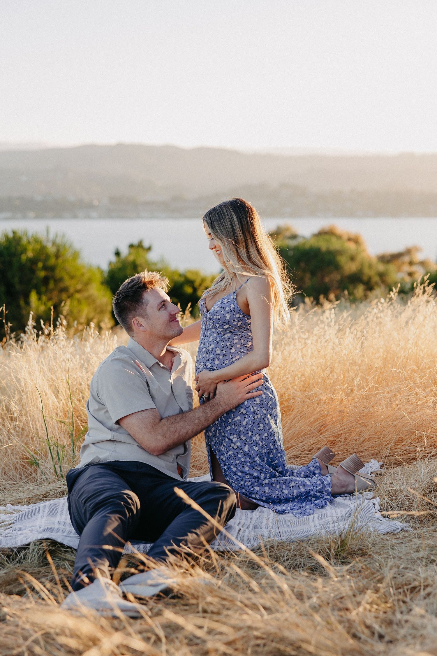 Romantic scene with couple sitting in tall grass with scenic lake and mountains in background.