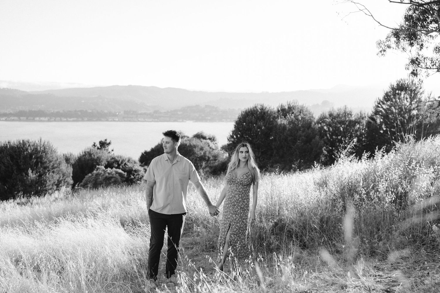 Black and white photo of couple in field holding hands with scenic backdrop.