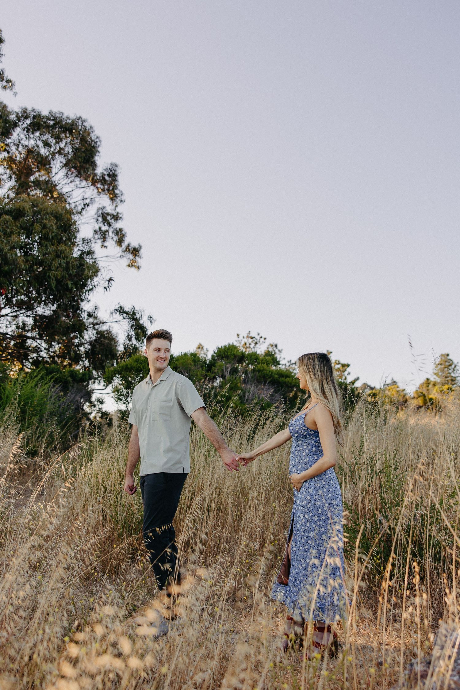 Couple holds hands while walking through golden dry grass with mountains in background.