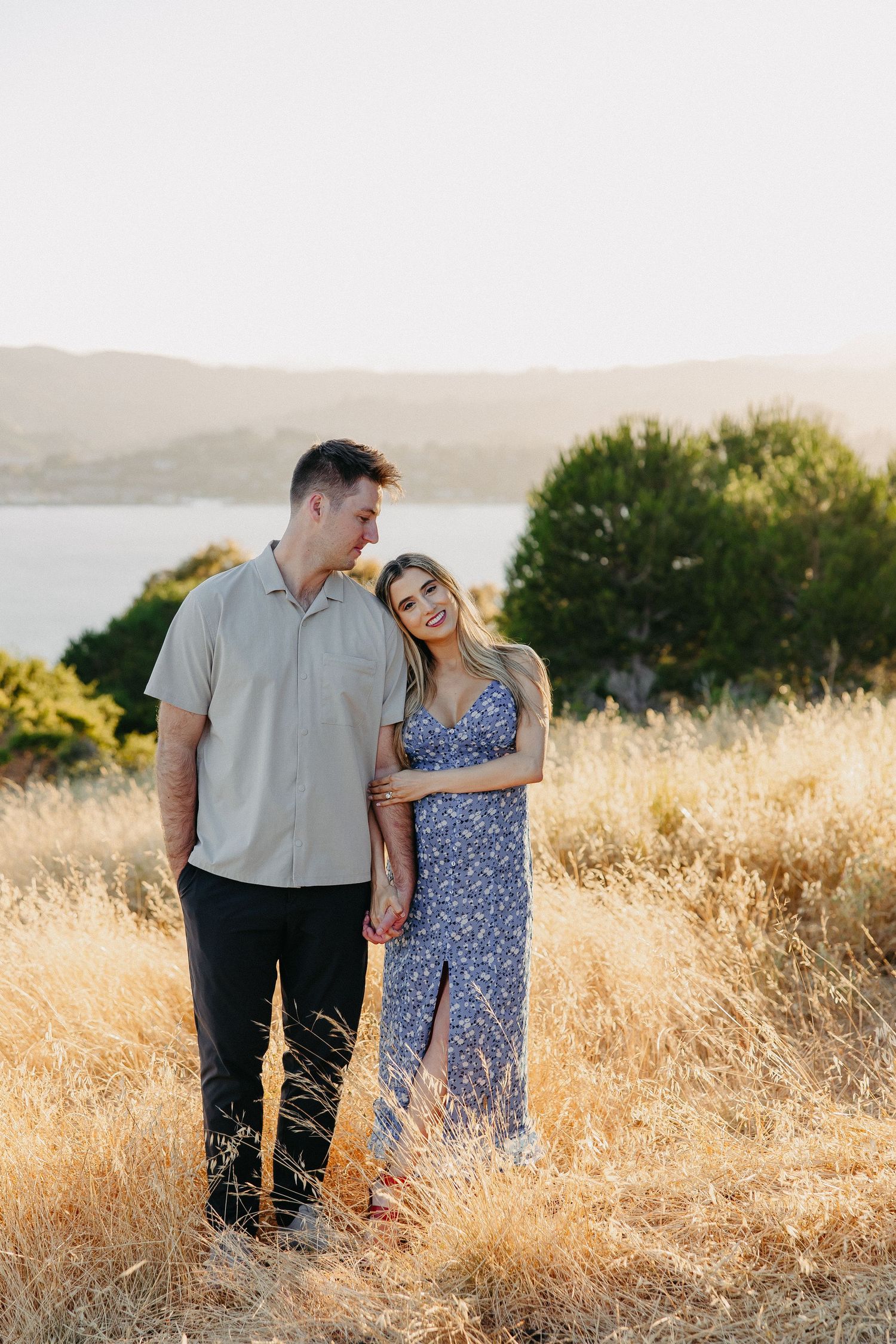 Happy couple standing together in golden field with lake view.