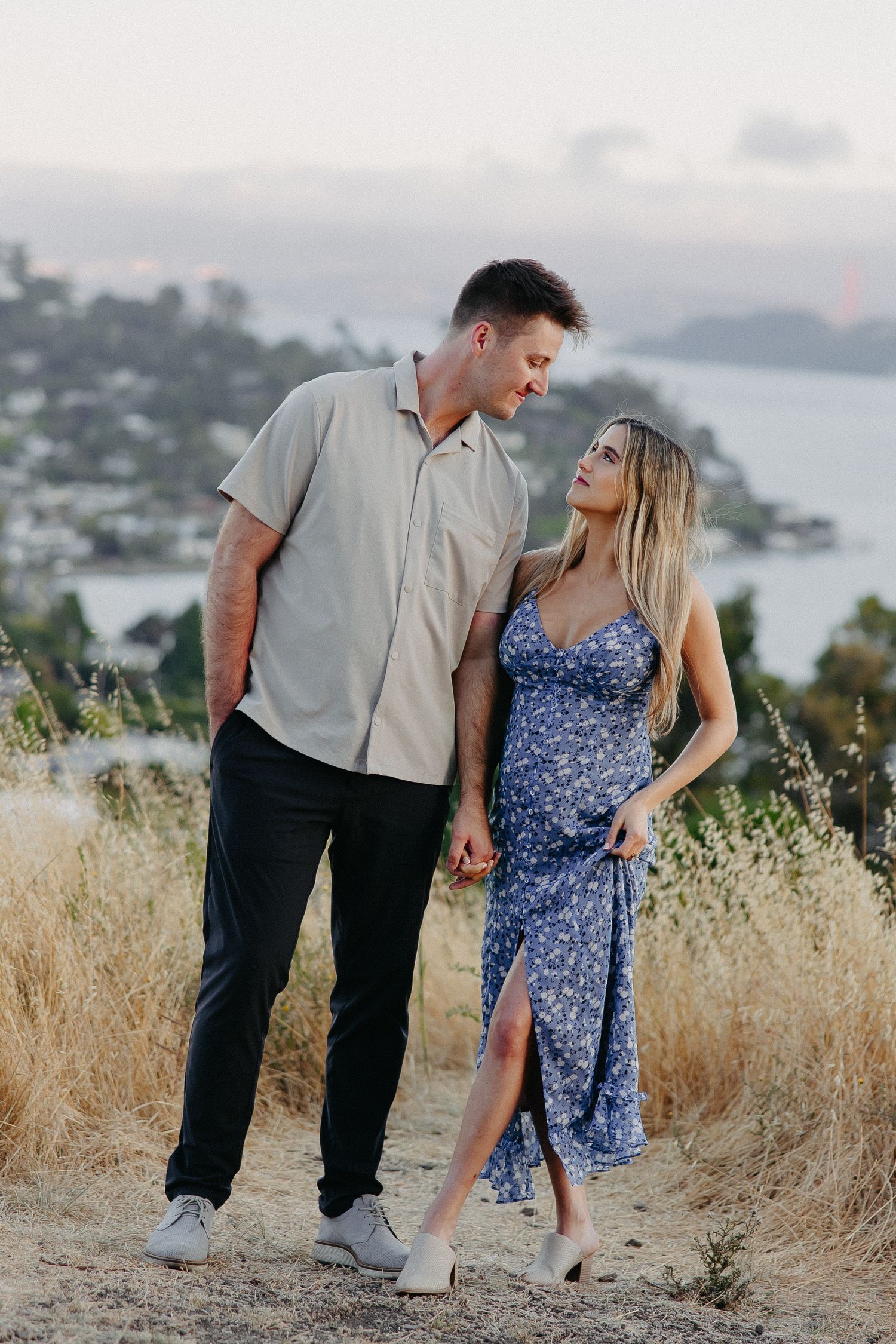 Floral dress and casual shirt clad couple pose together on scenic overlook with bay views.