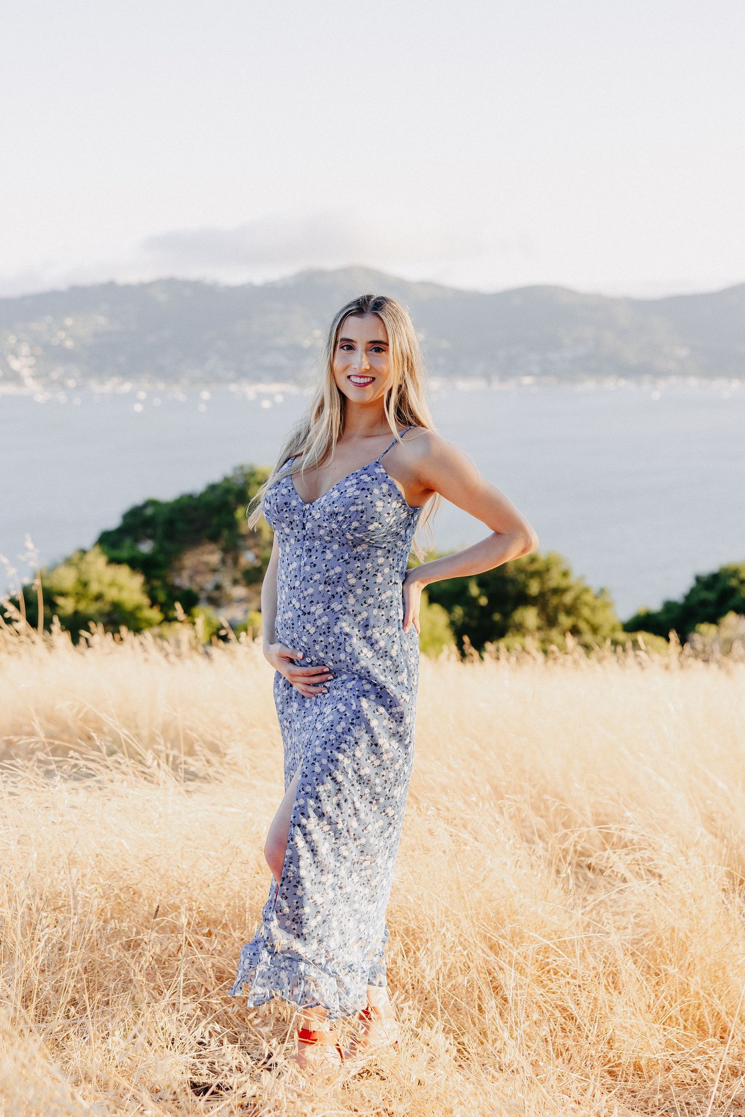 Expectant woman in blue floral maxi dress poses against mountainous coastal backdrop.