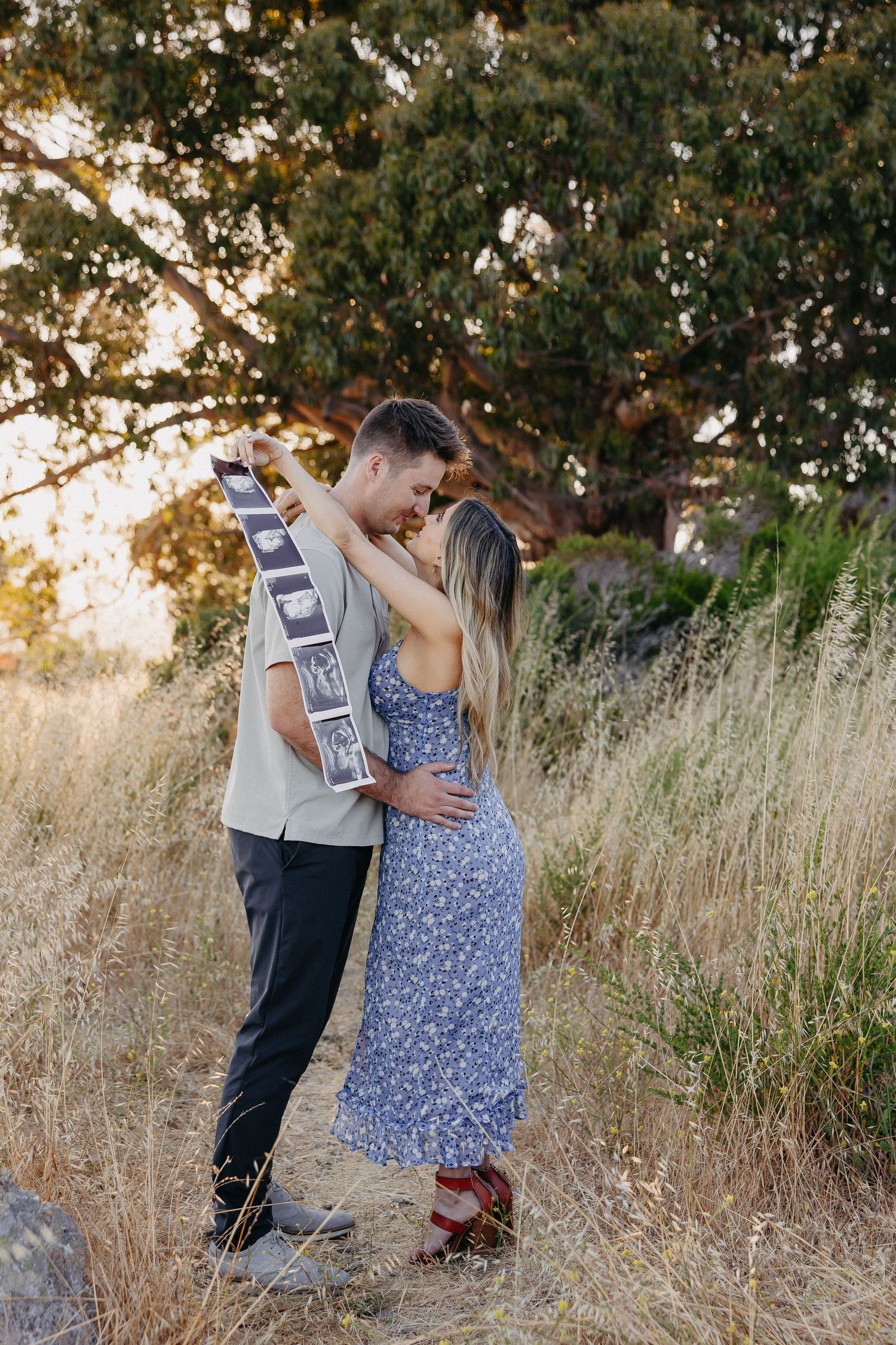 Couple holding ultrasound photos while embracing in sunlit field with trees in background.