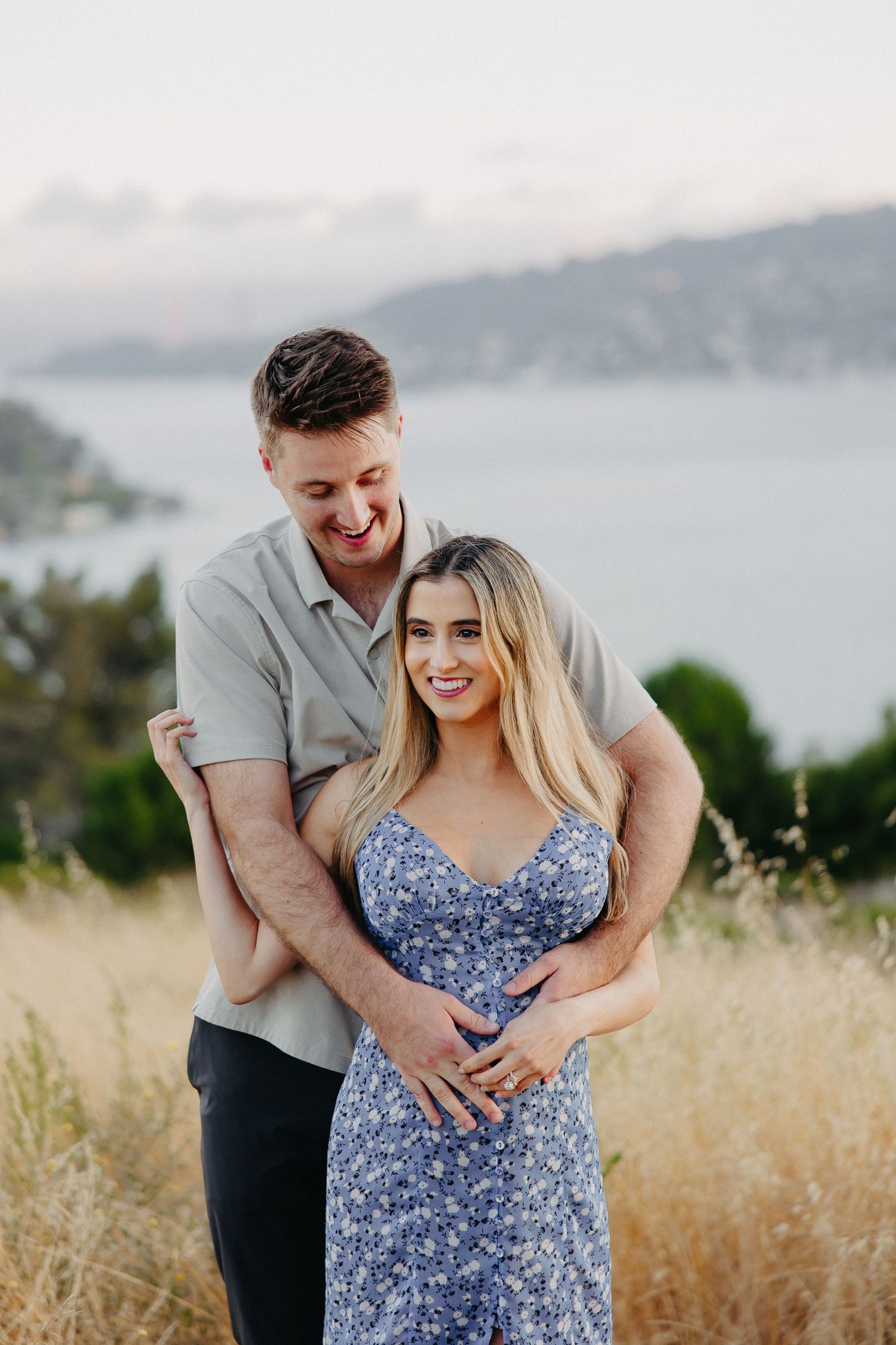 Loving couple sharing intimate embrace on hillside with water and mountains in distance.