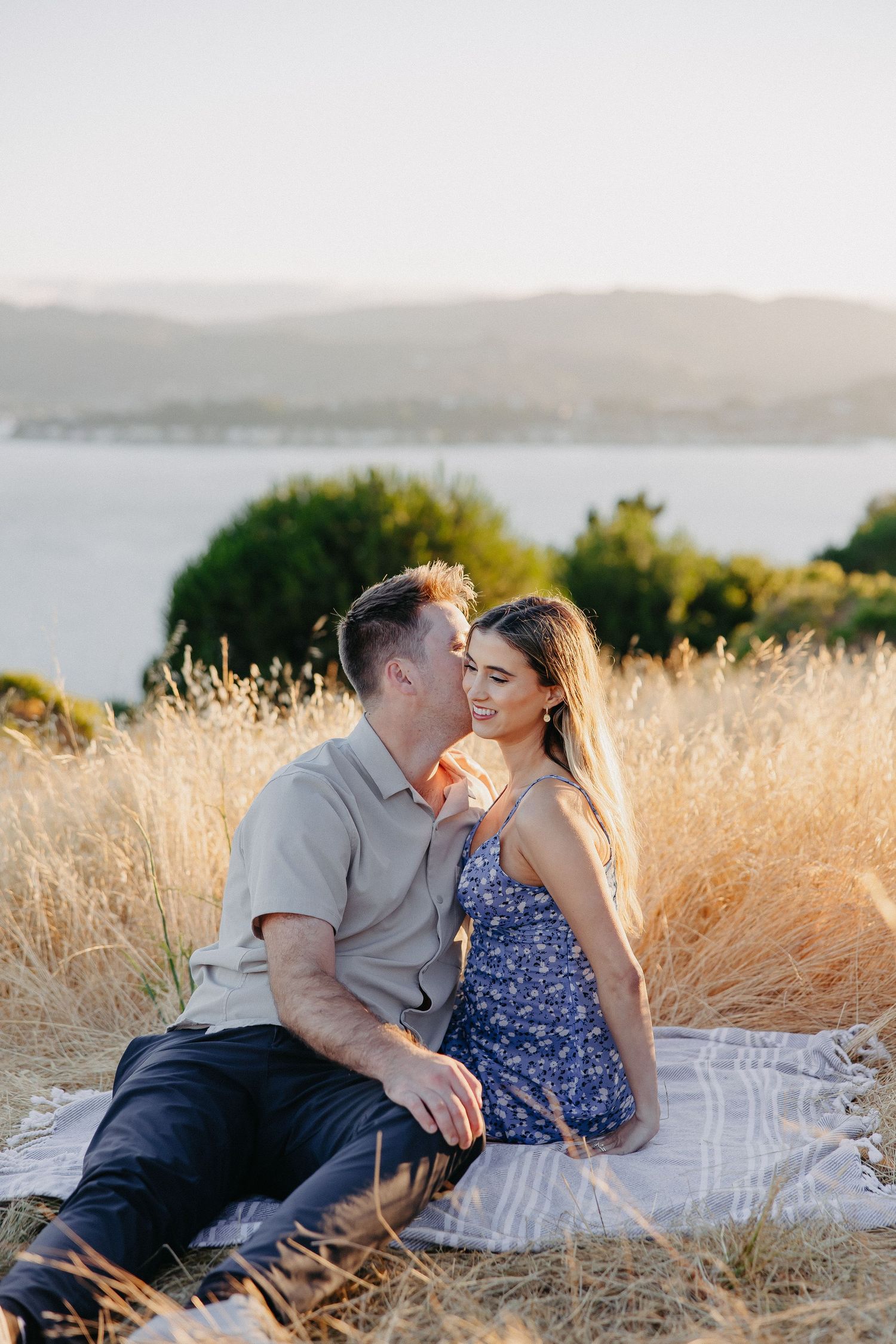 Tender moment between couple sitting on blanket in sunlit field with water view.