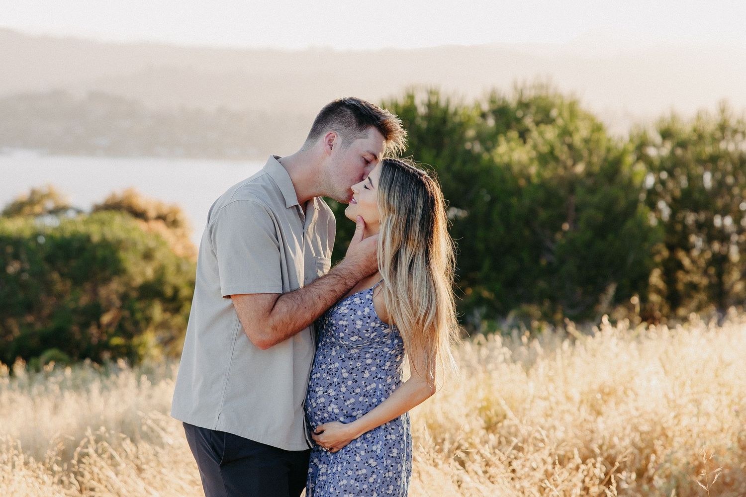 A couple embracing in golden meadow at sunset with water and mountains in background.