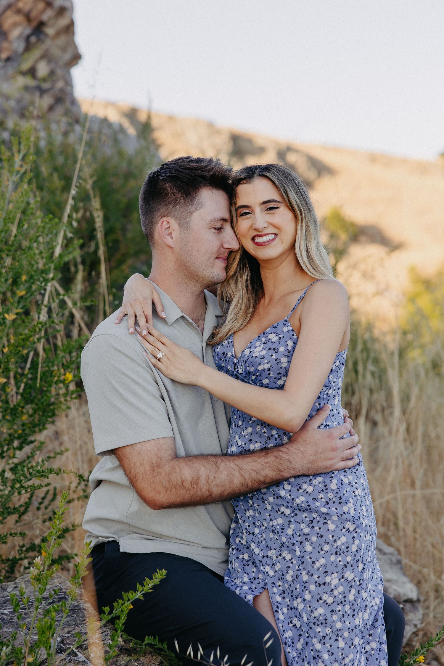 A couple embraces in golden sunlit field wearing blue floral dress and beige shirt against mountain backdrop.