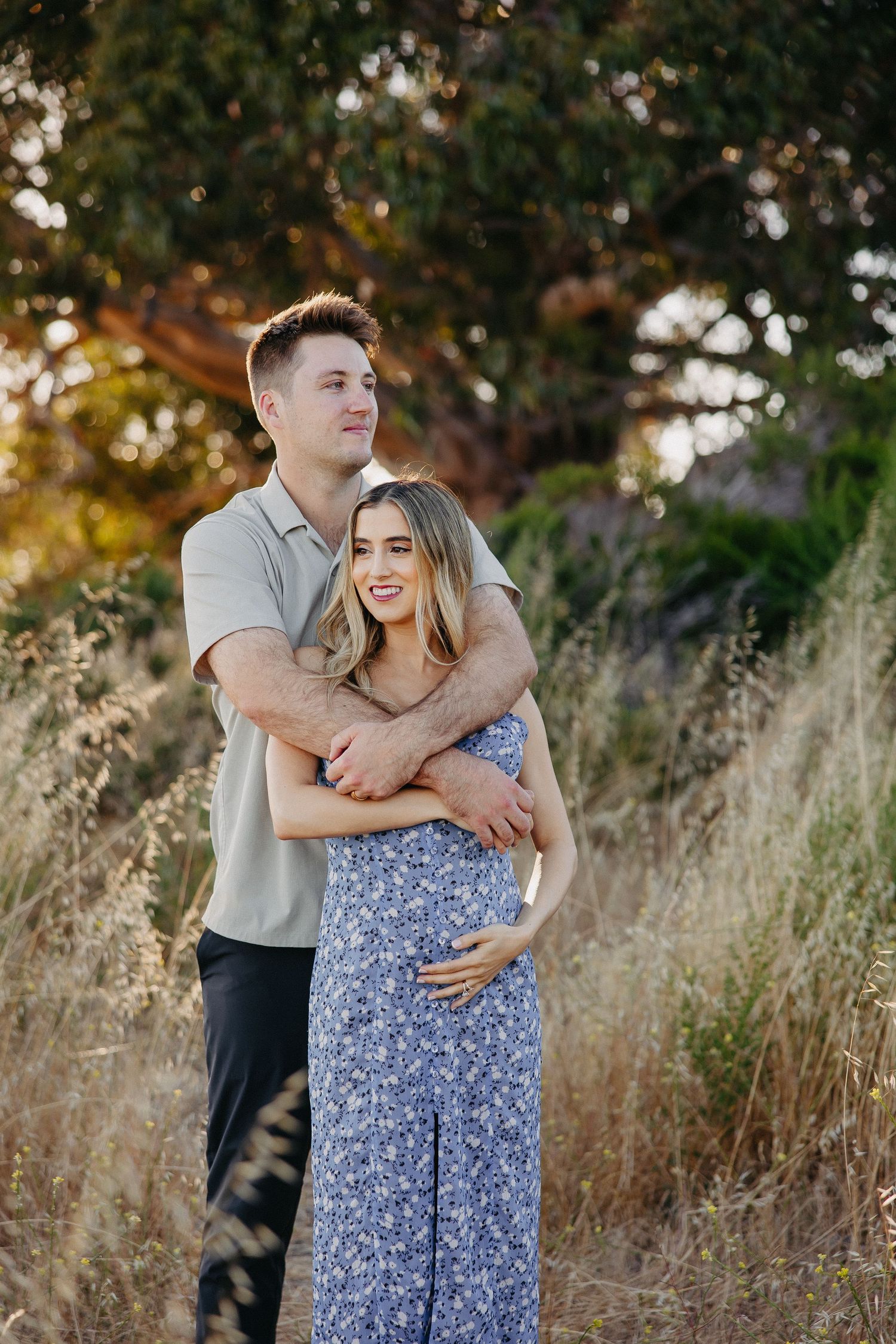 Couple embracing in golden sunlight with tall grass and trees in background.