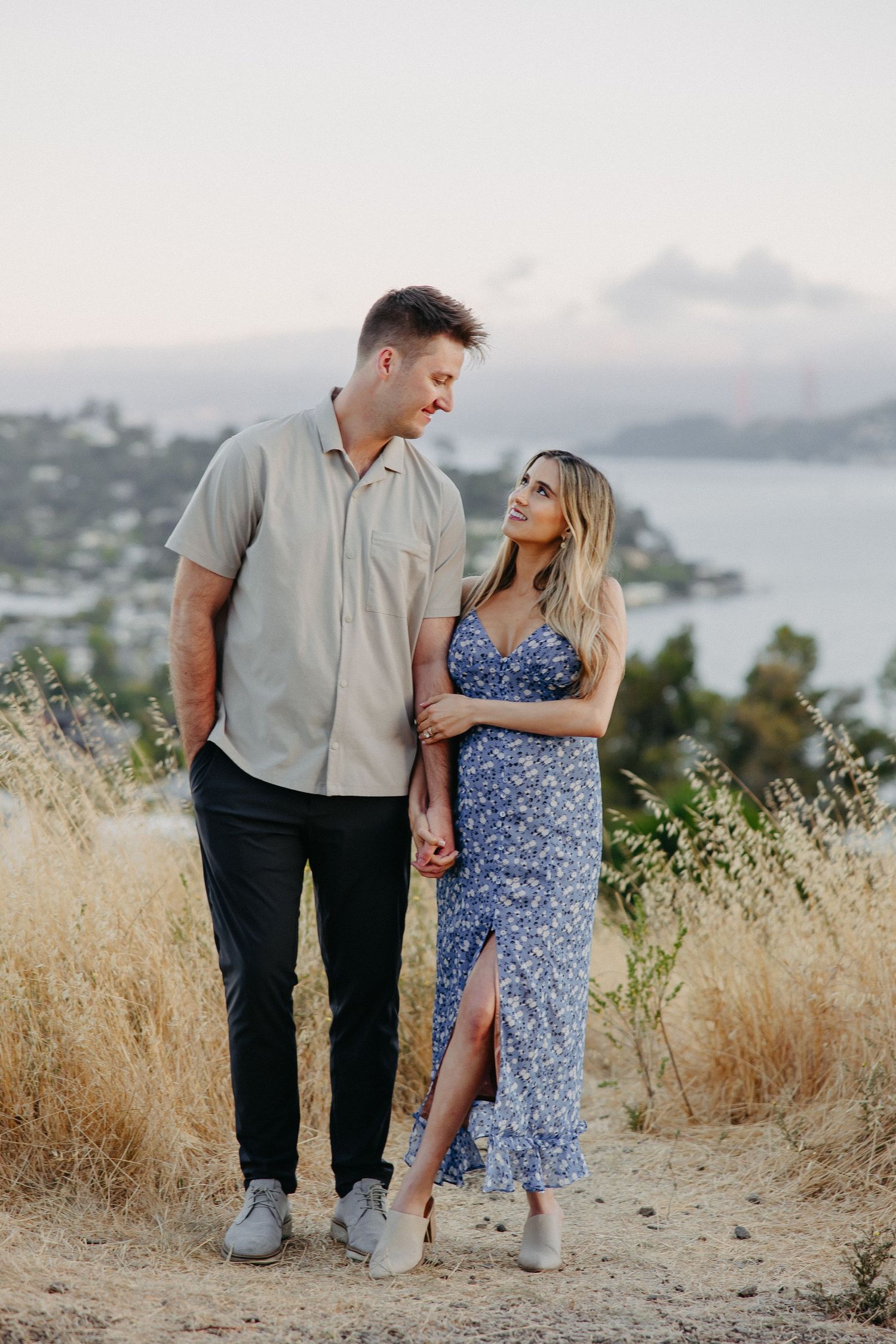 Couple in casual attire sharing a romantic moment on hilltop overlooking water and mountains at sunset.
