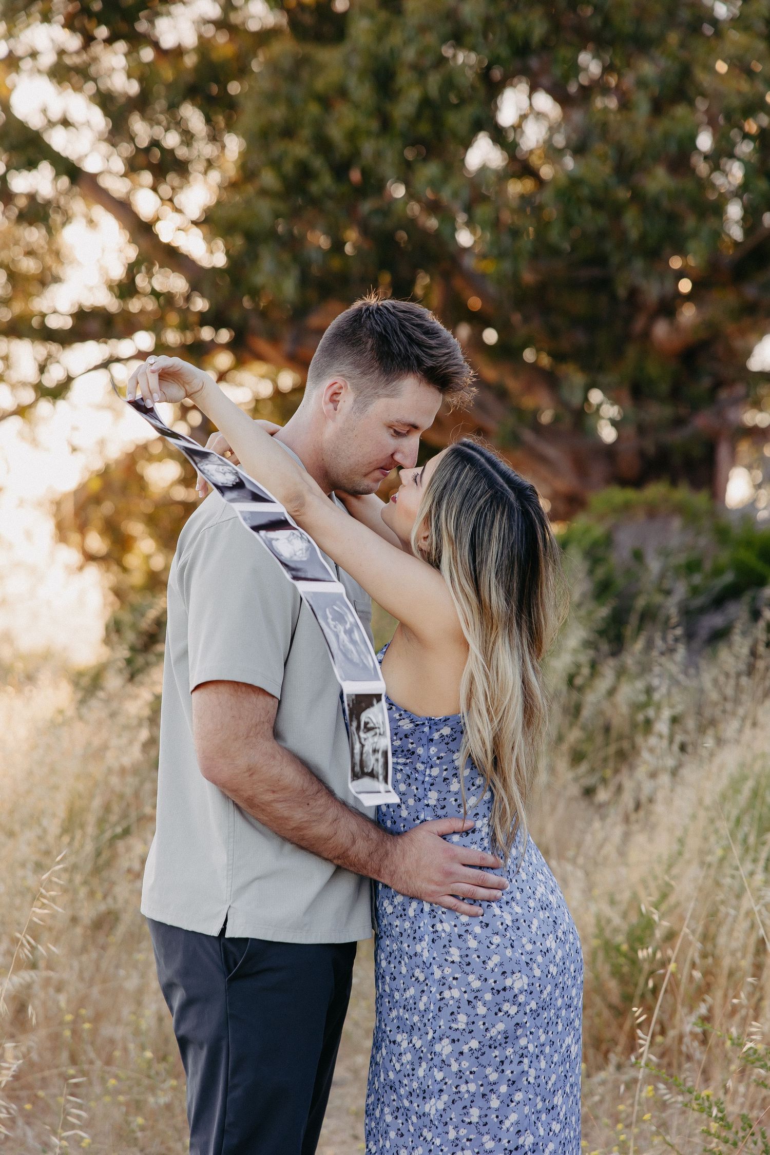 Intimate moment between couple in natural setting while holding pregnancy announcement photos.