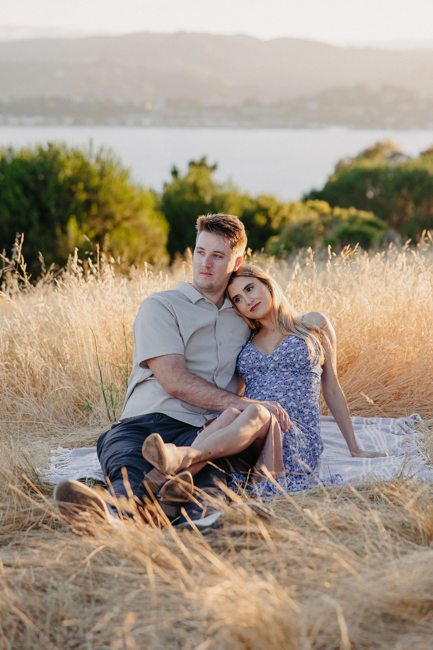 Couple relaxing together on blanket in golden field with scenic water backdrop.