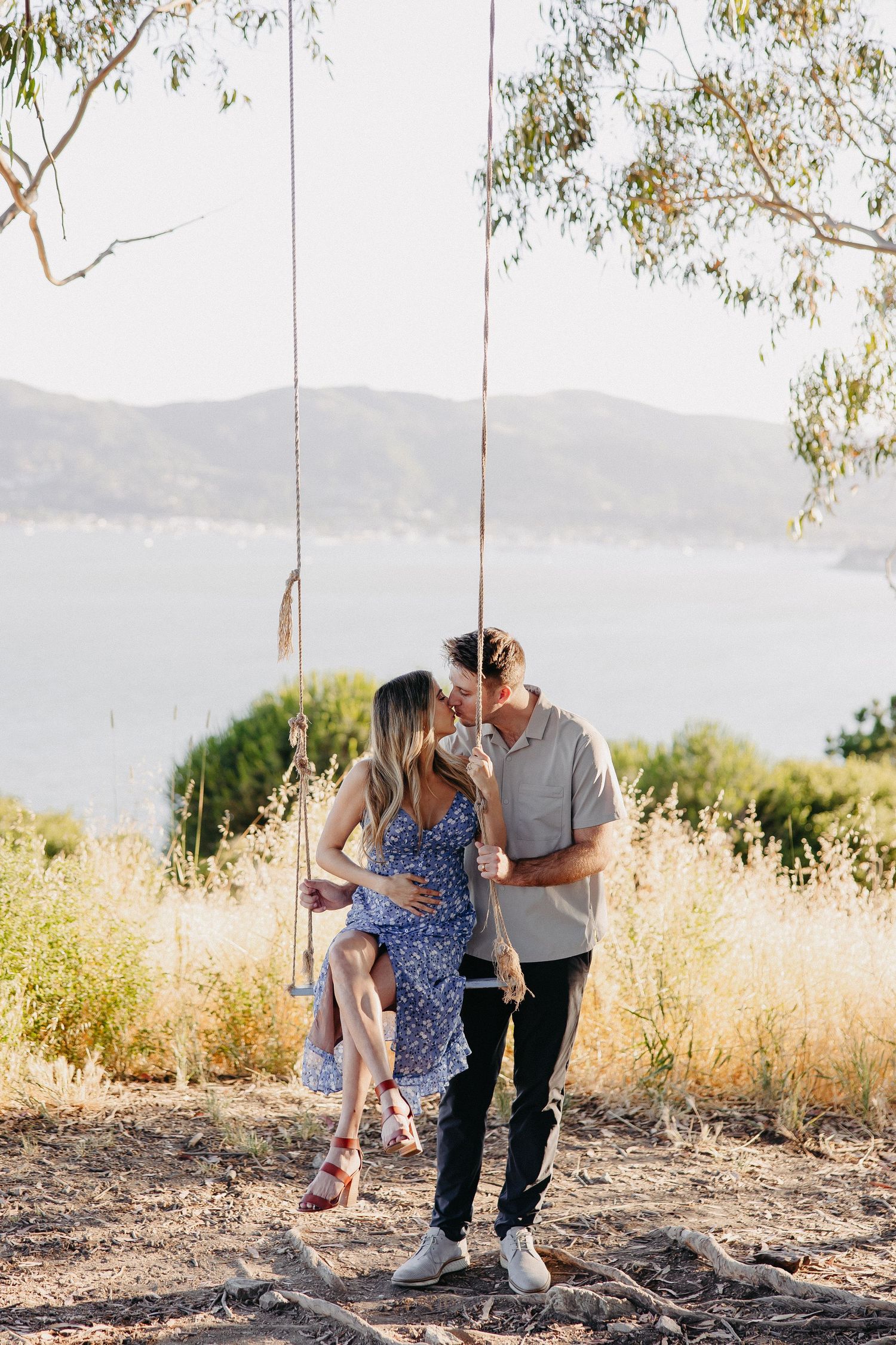 A couple shares a romantic moment on a tree swing overlooking a serene mountain landscape.