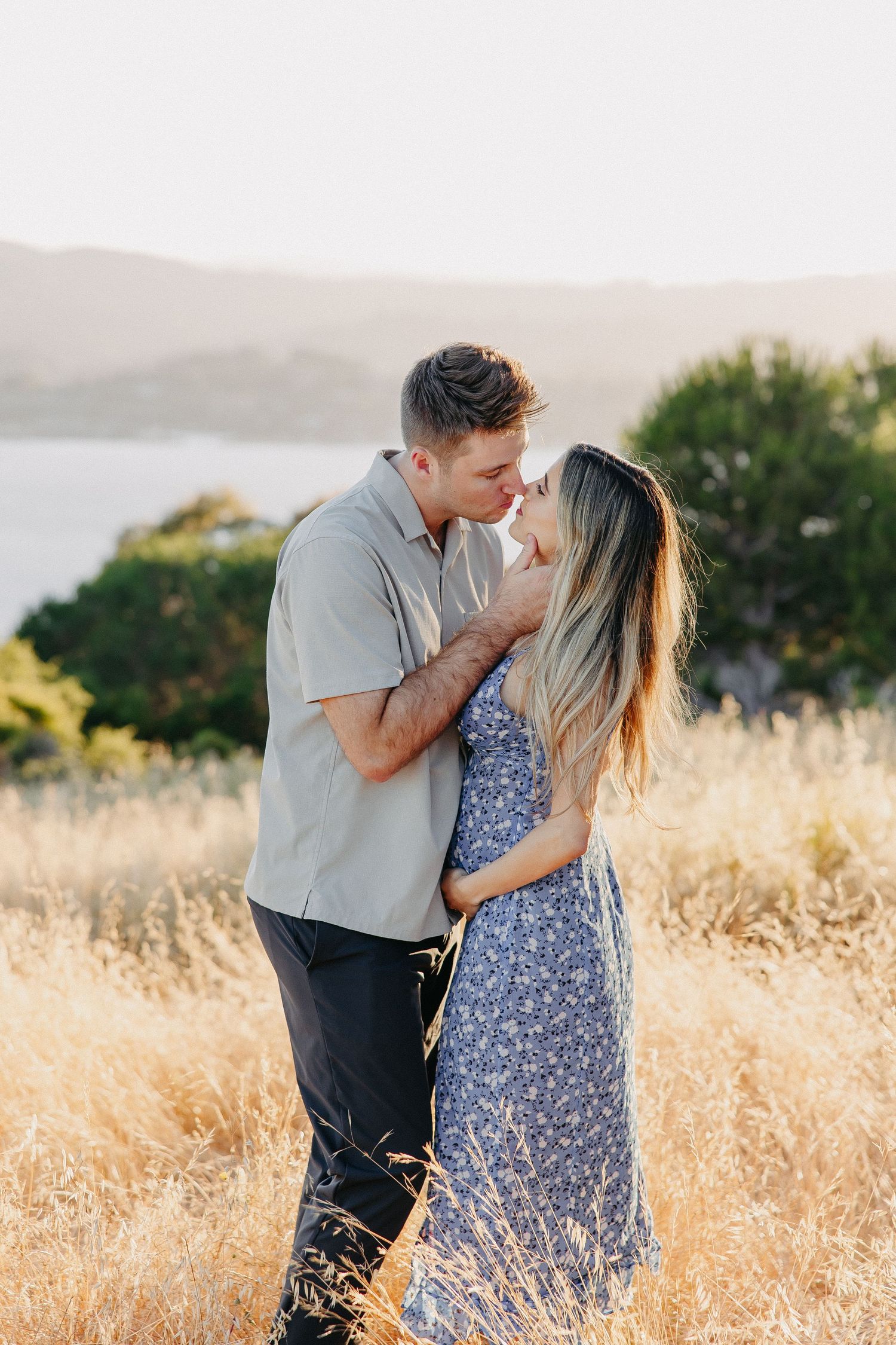 Tender moment between couple in golden field with water view during sunset maternity shoot.