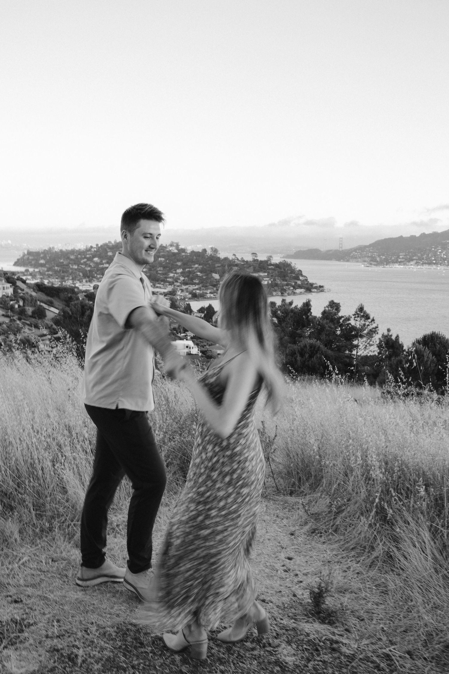 Black and white photo of couple dancing in field overlooking bay.