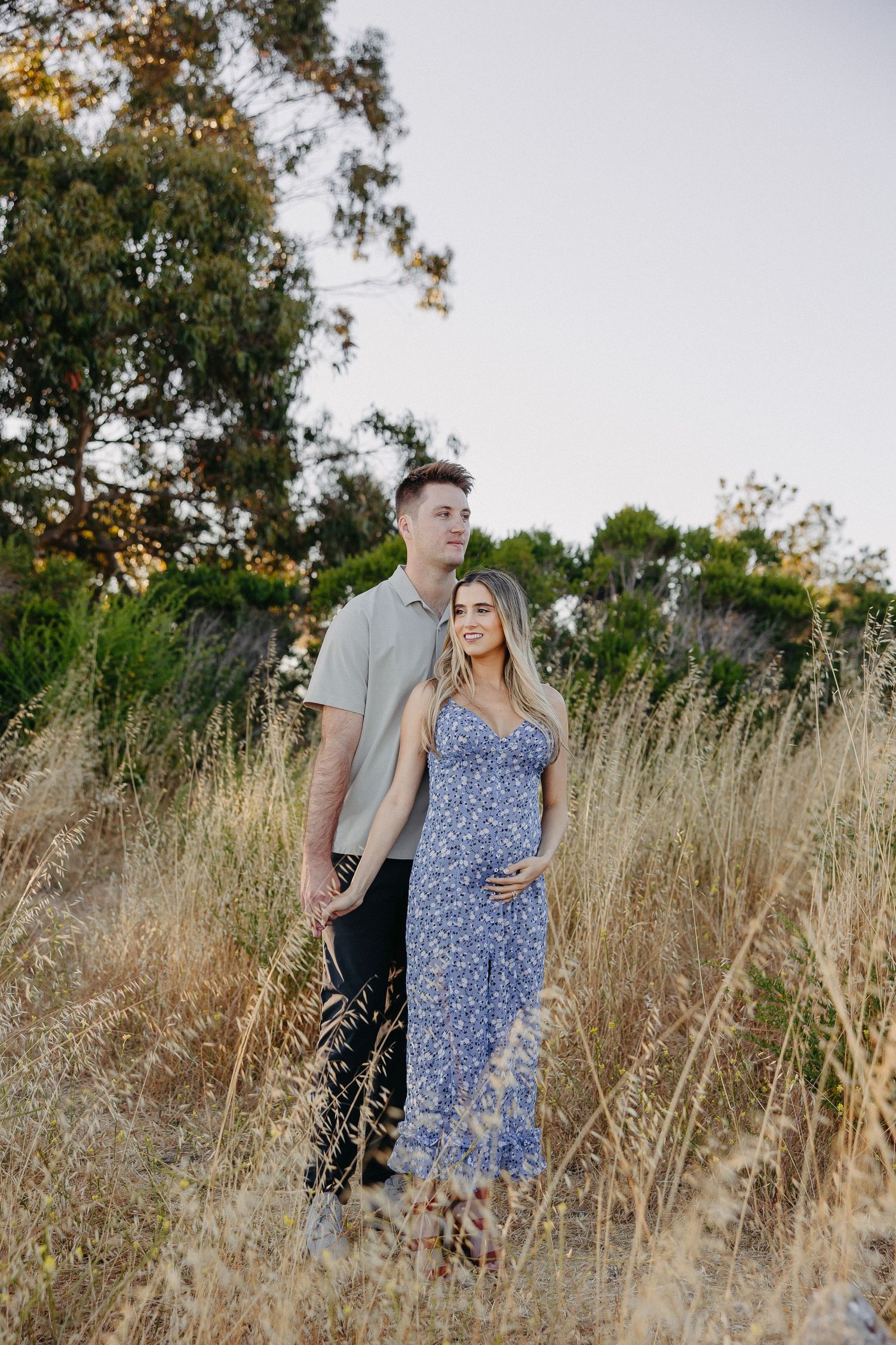 Couple stands together in field of tall grass during sunset photoshoot.