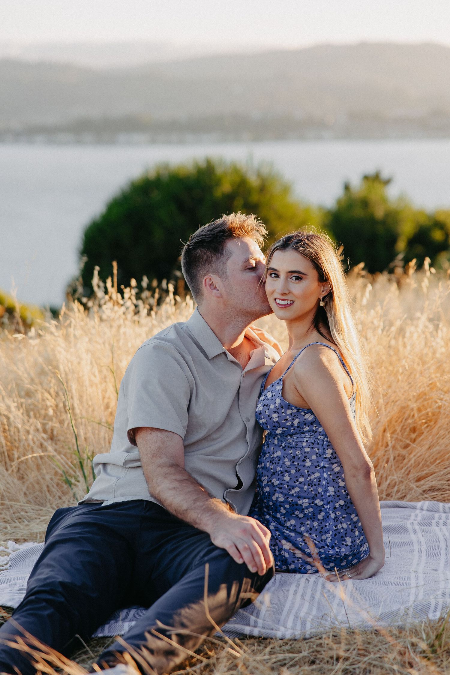 Couple sharing affectionate moment on hillside during golden hour with lake vista.