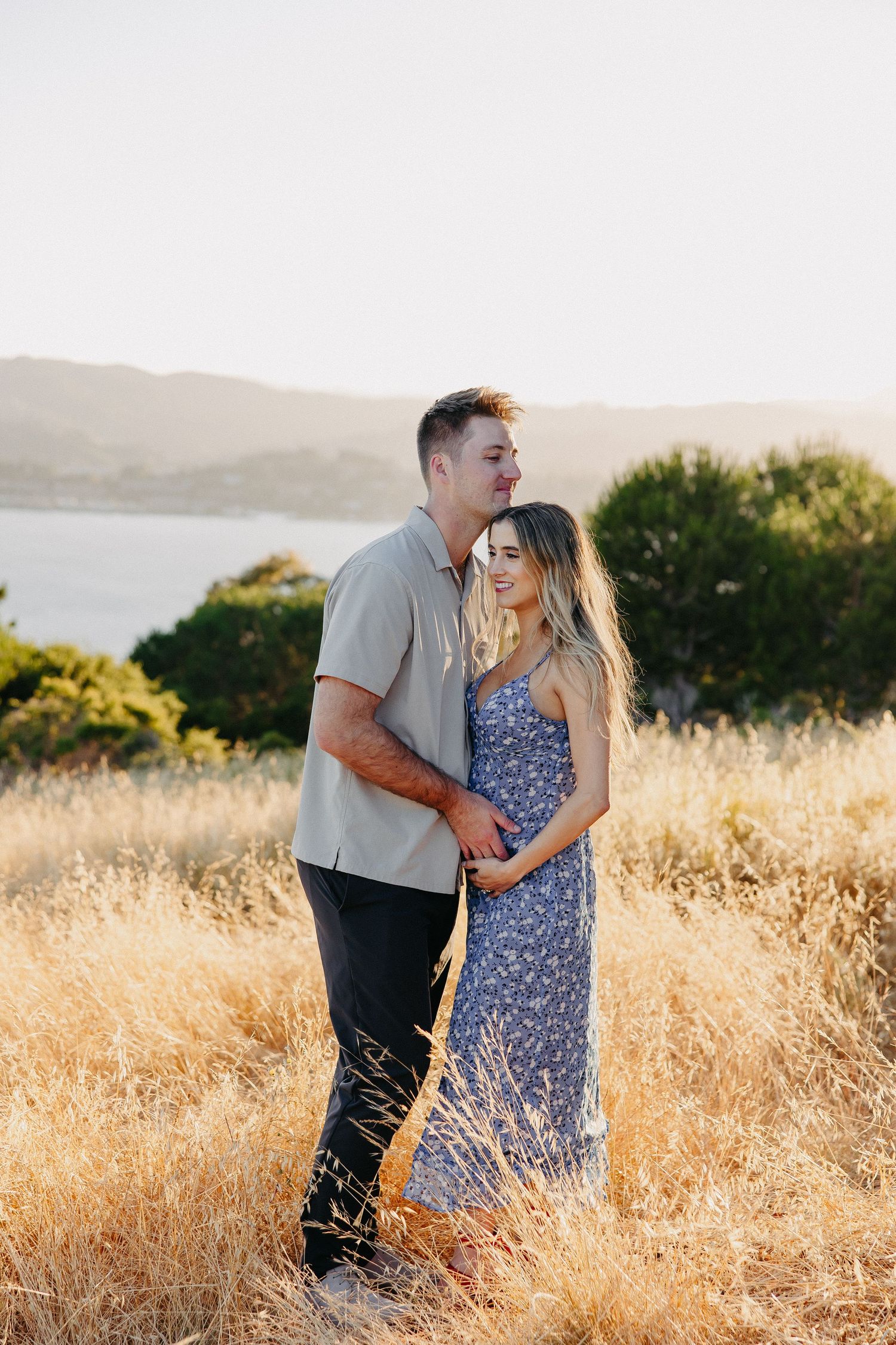 Couple sharing tender moment in sunlit field overlooking water.