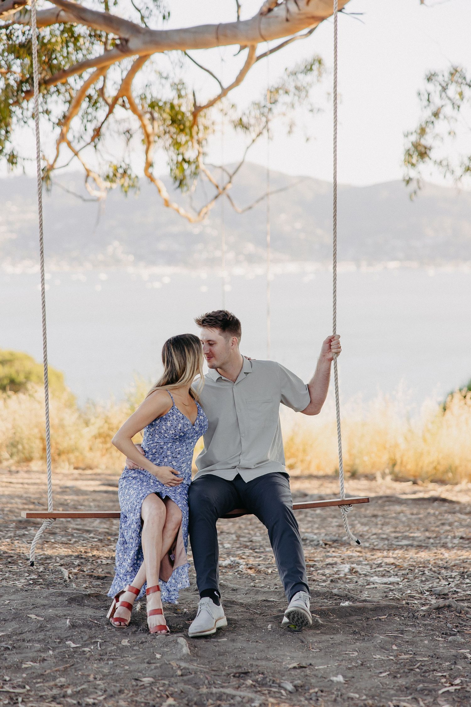 Couple sits together on a rustic swing with mountains and lake in the background.