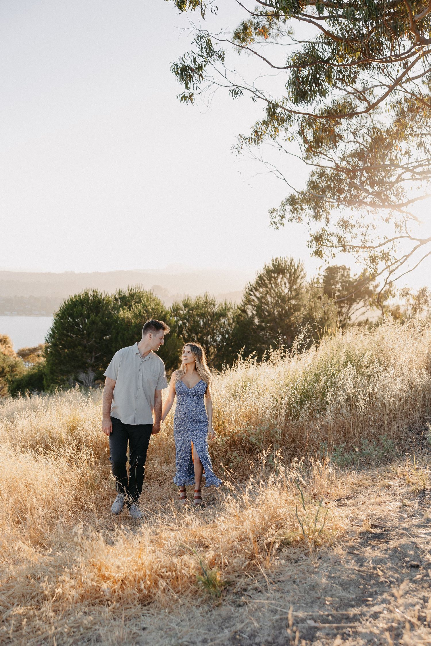Couple walking hand in hand through golden grass with water view.