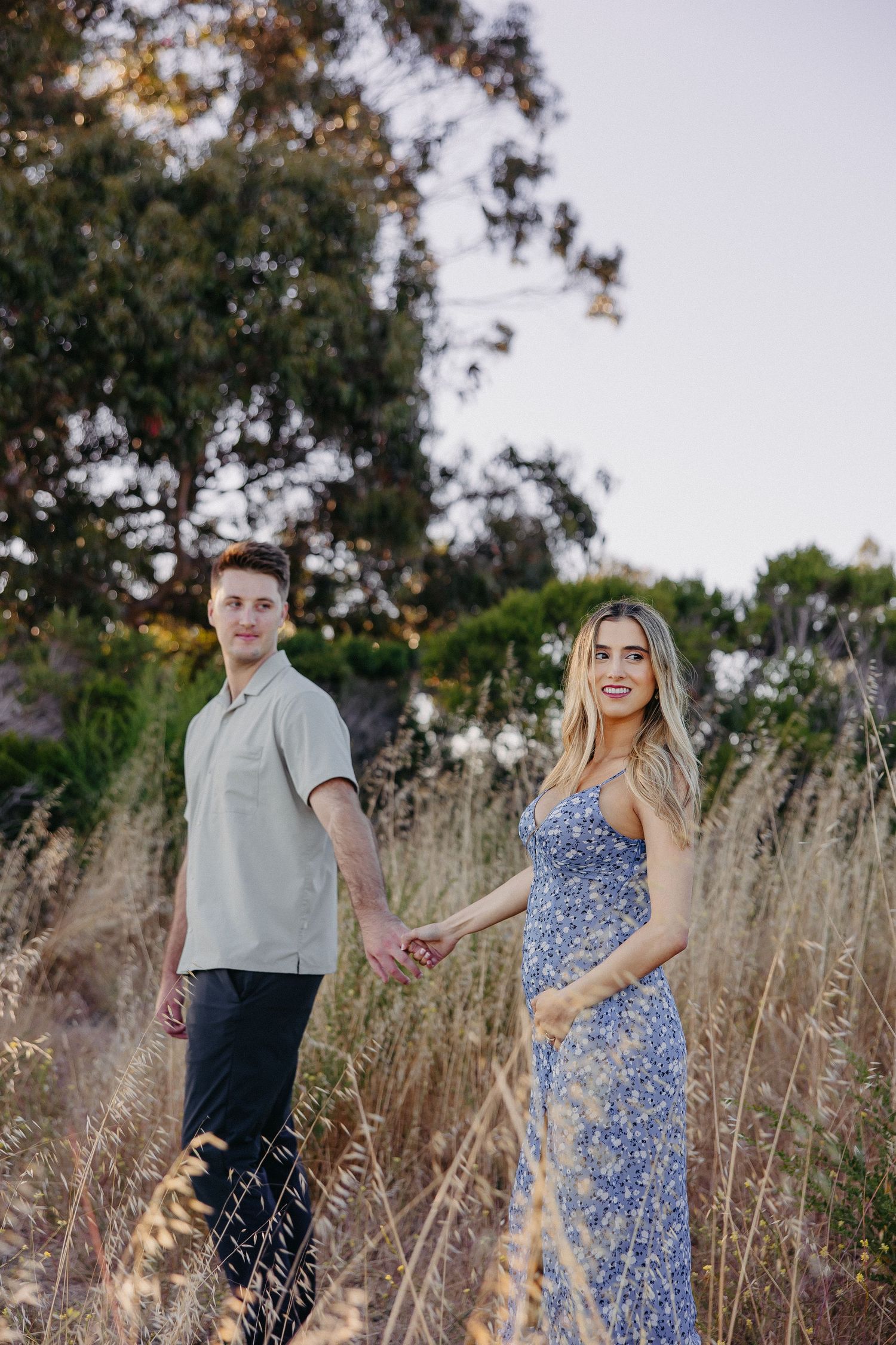 Couple poses in a field of tall grass during golden hour with trees behind them.
