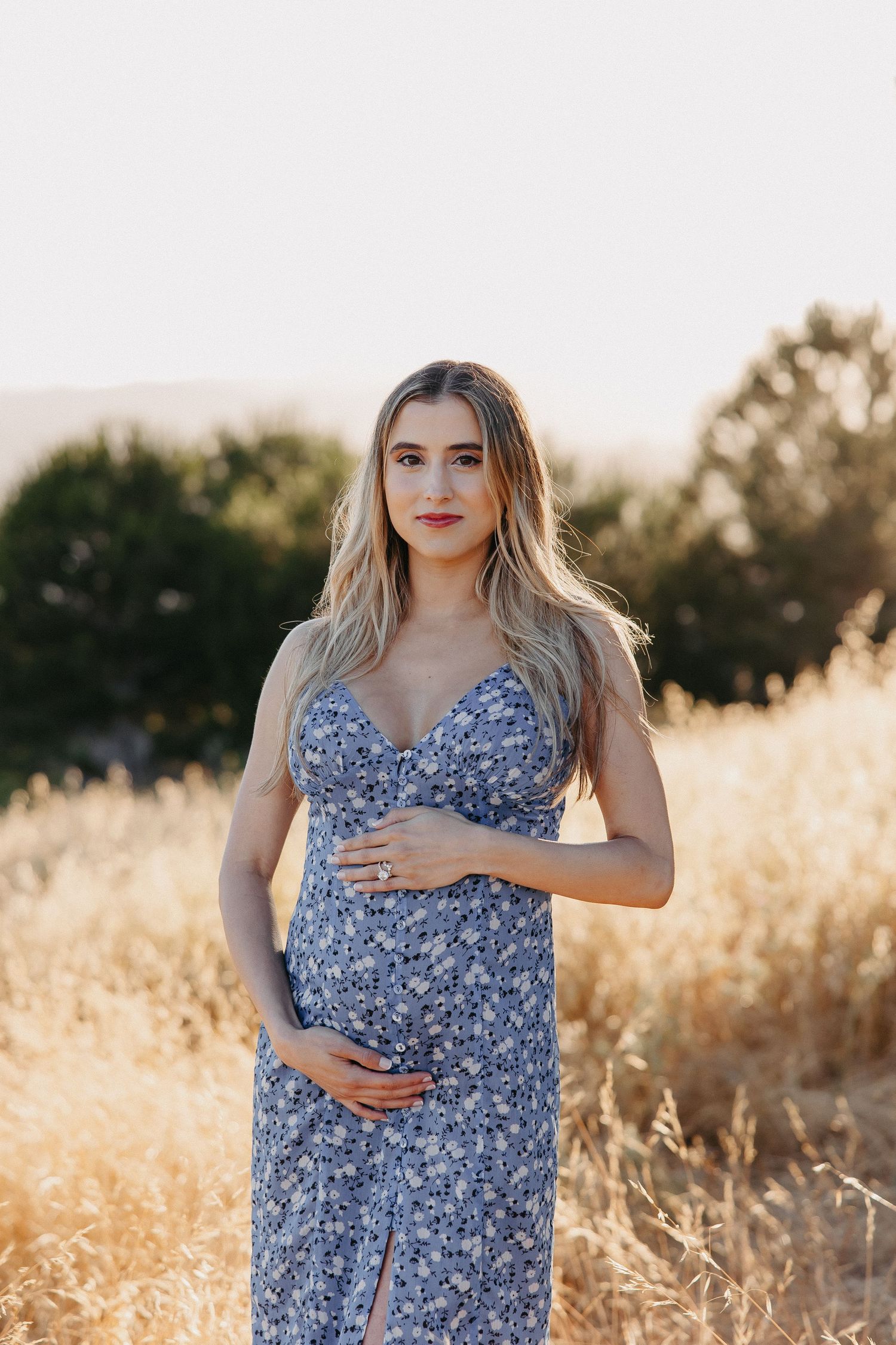 Expectant mother in blue floral dress poses against scenic hillside background.