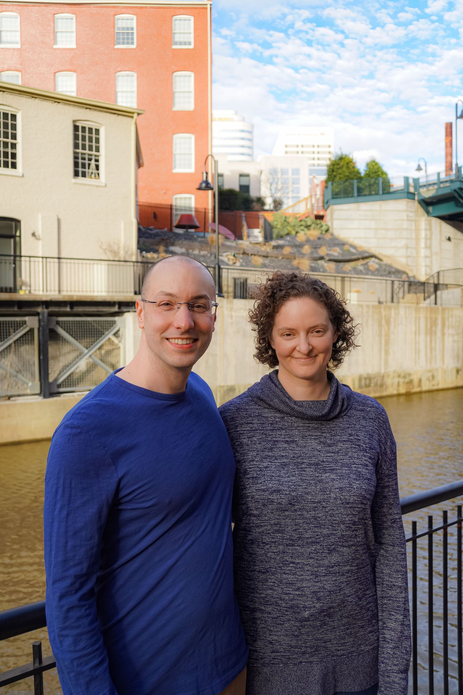 Couple posing together by a canal with brick buildings and city skyline in background.