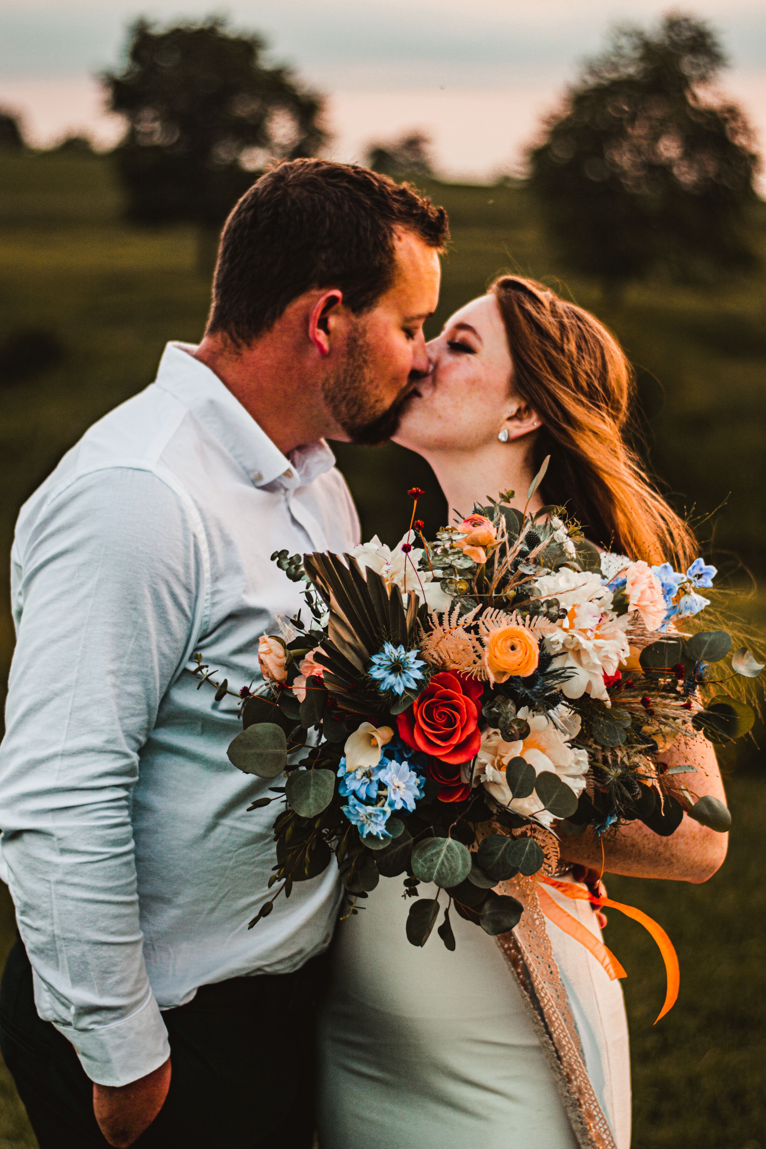 Tender moment between newlyweds in a field holding vibrant floral bouquet at sunset.