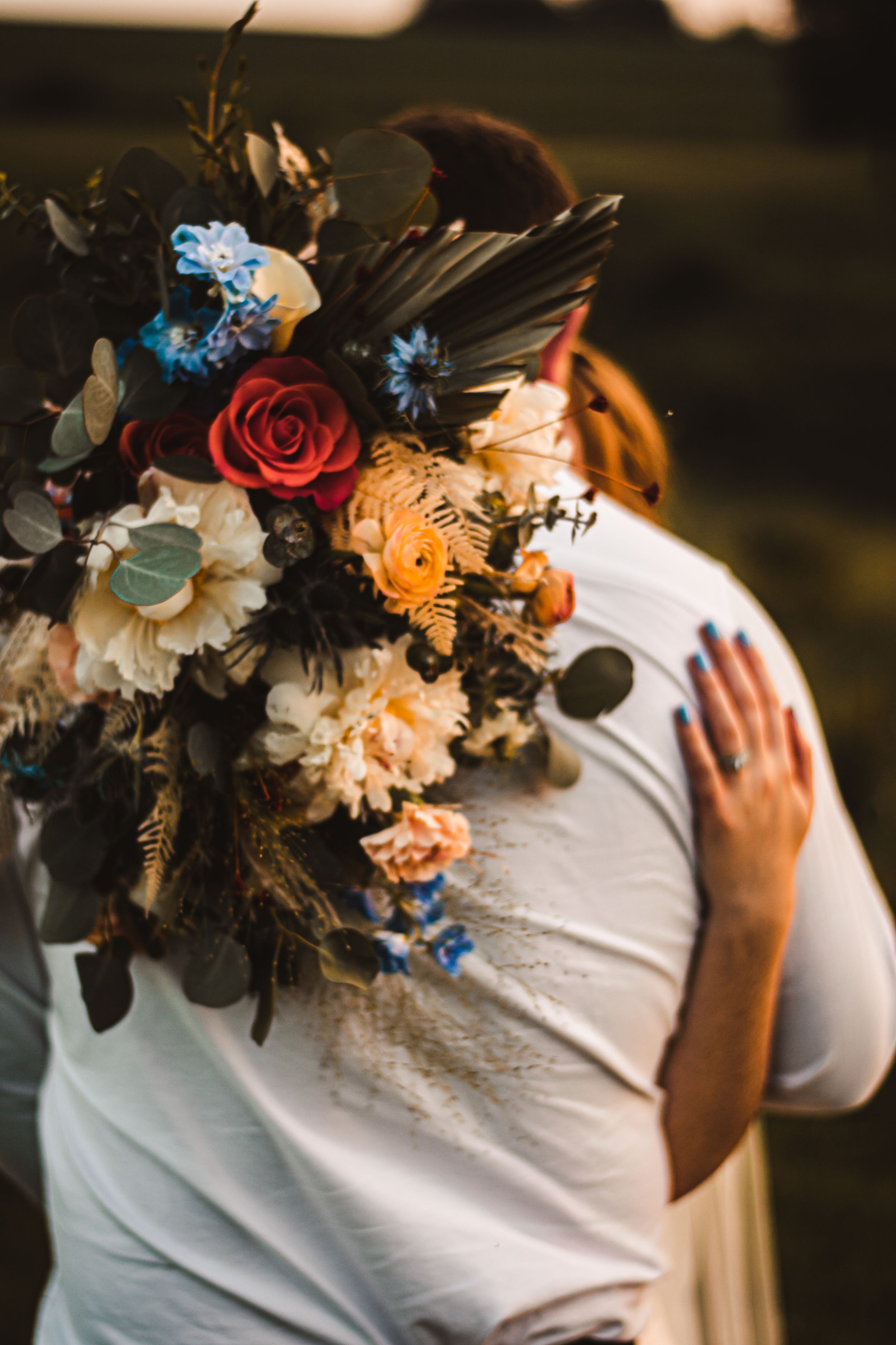 A close-up of a wedding bouquet with red roses, blue flowers, and feathers against white fabric.