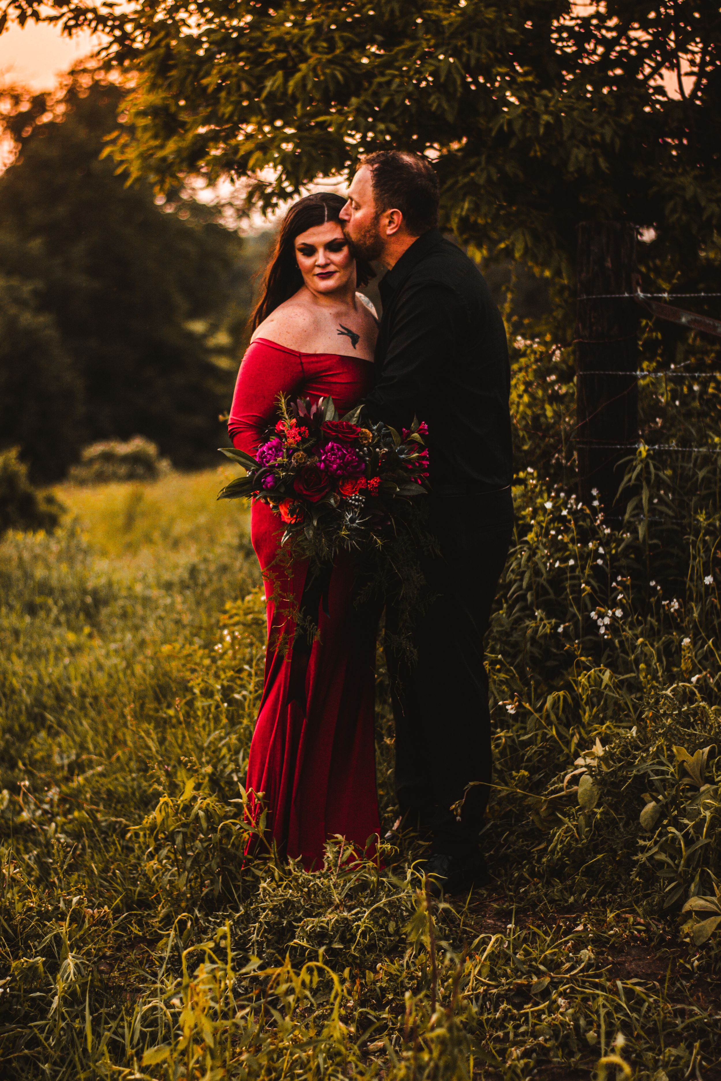 Sunset engagement photo with dramatic red dress and lush bouquet in natural setting.