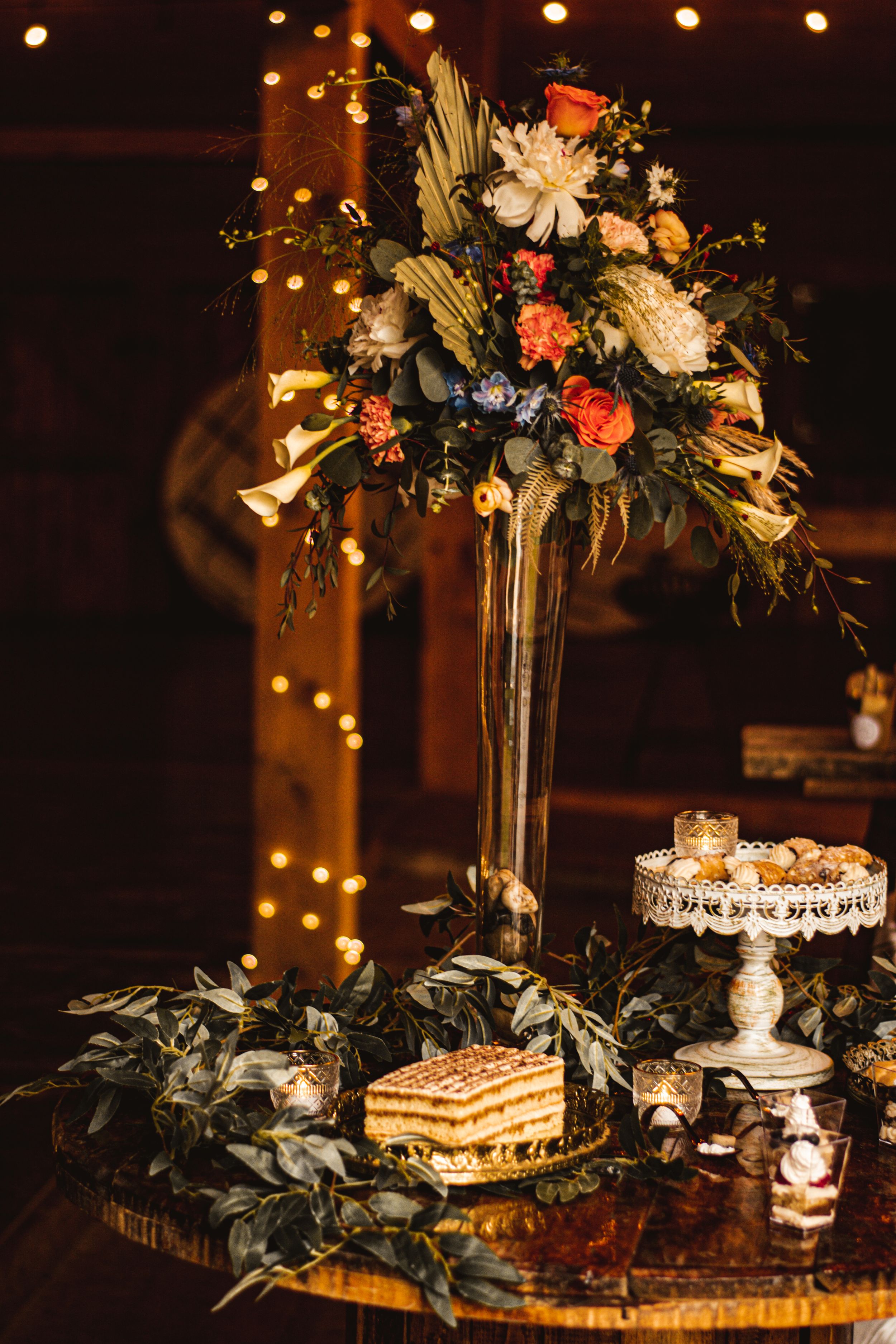 Elegant wedding reception display with tall floral centerpiece and dessert table in moody lighting.