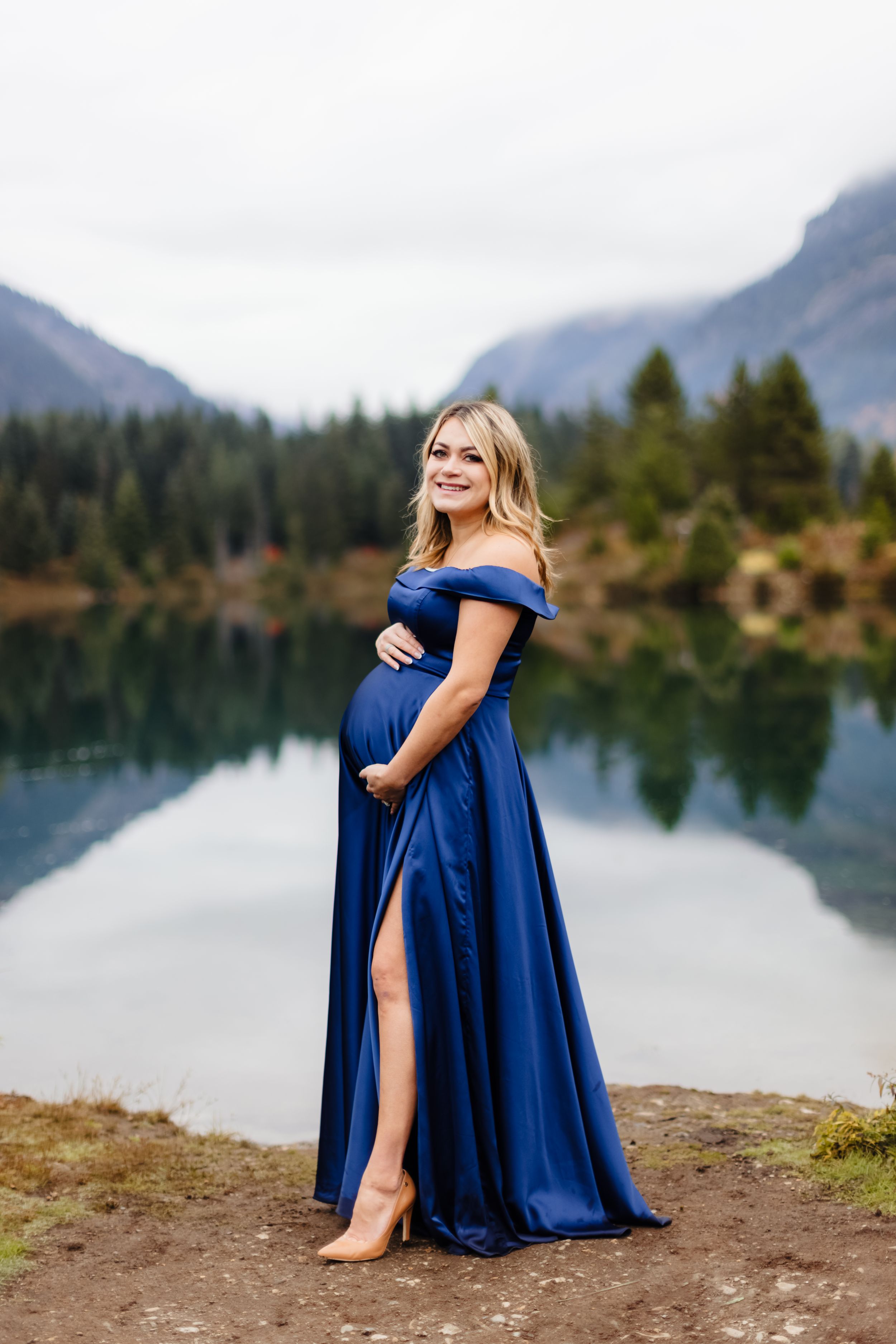 A pregnant woman in a flowing blue gown poses by a scenic mountain lake.