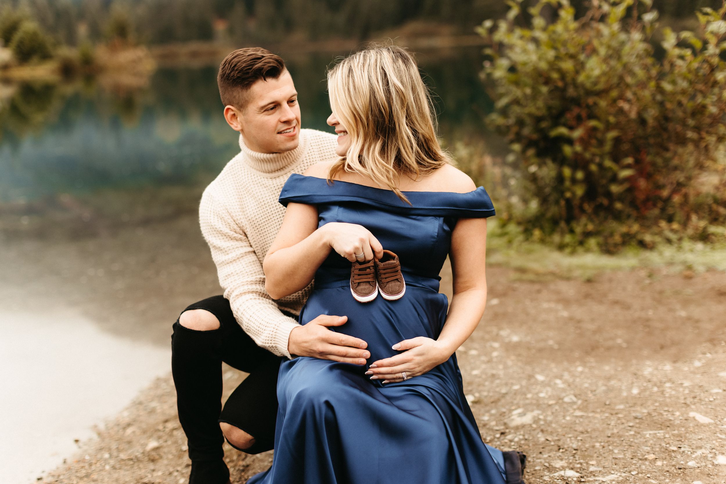 A couple poses together by a lake with mountains in the background.
