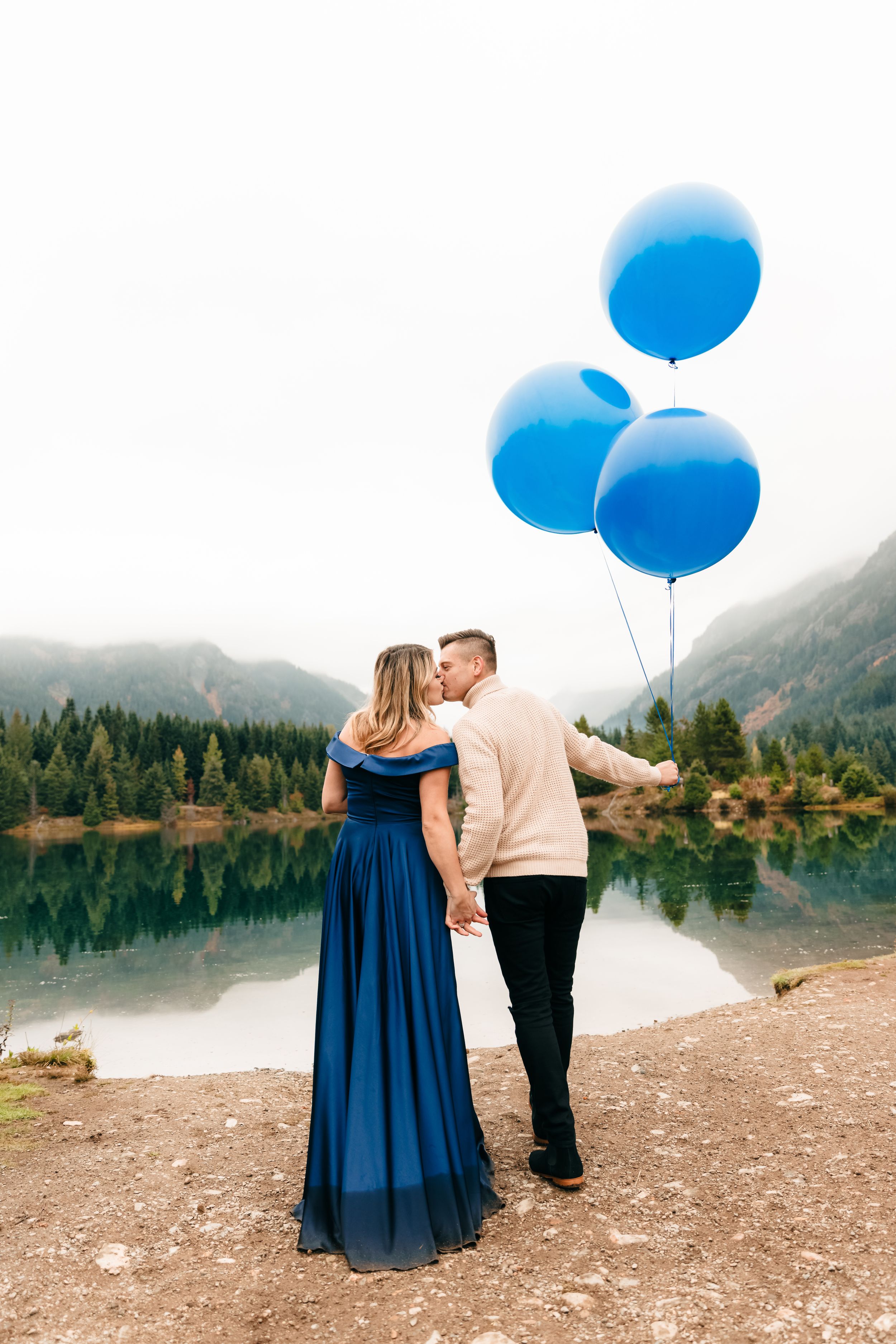 A couple holds blue balloons while standing by a scenic mountain lake.