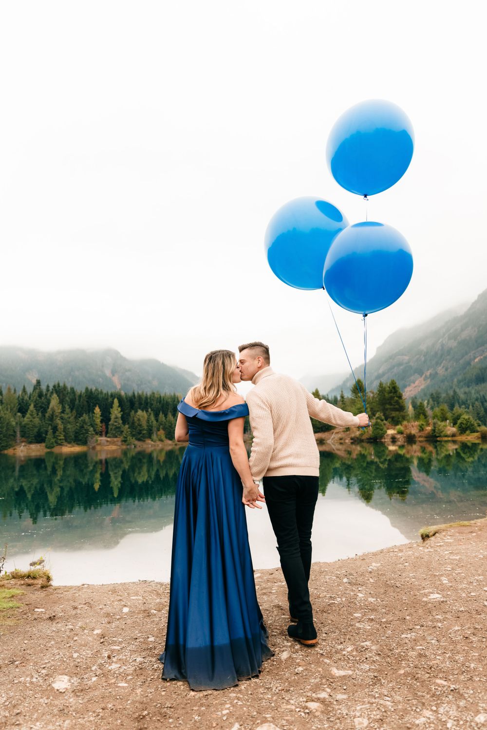 A couple holds blue balloons while standing by a scenic mountain lake.