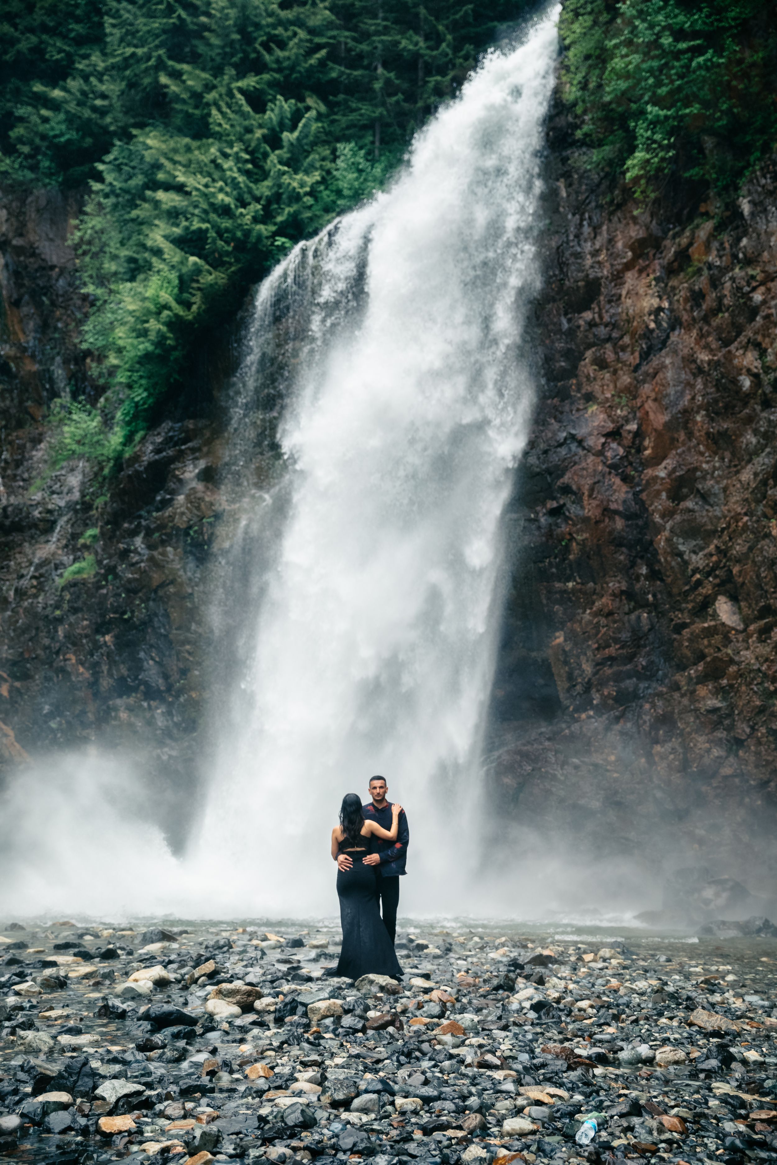 A couple embraces in front of a majestic waterfall surrounded by forest.