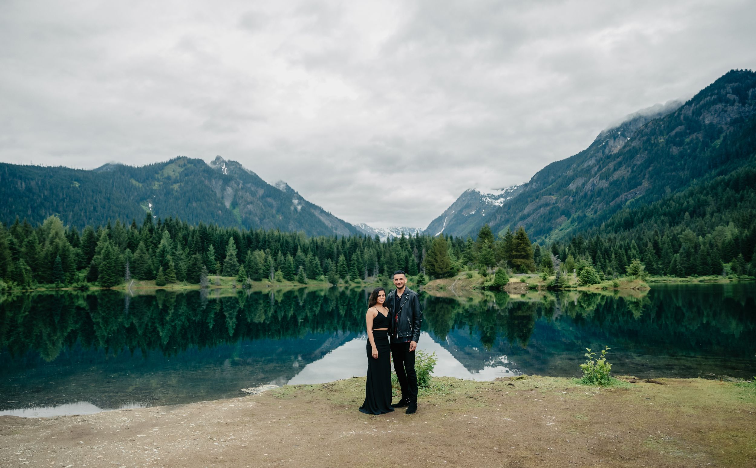 A couple stands on a rocky shore with mountains and a lake in the background.