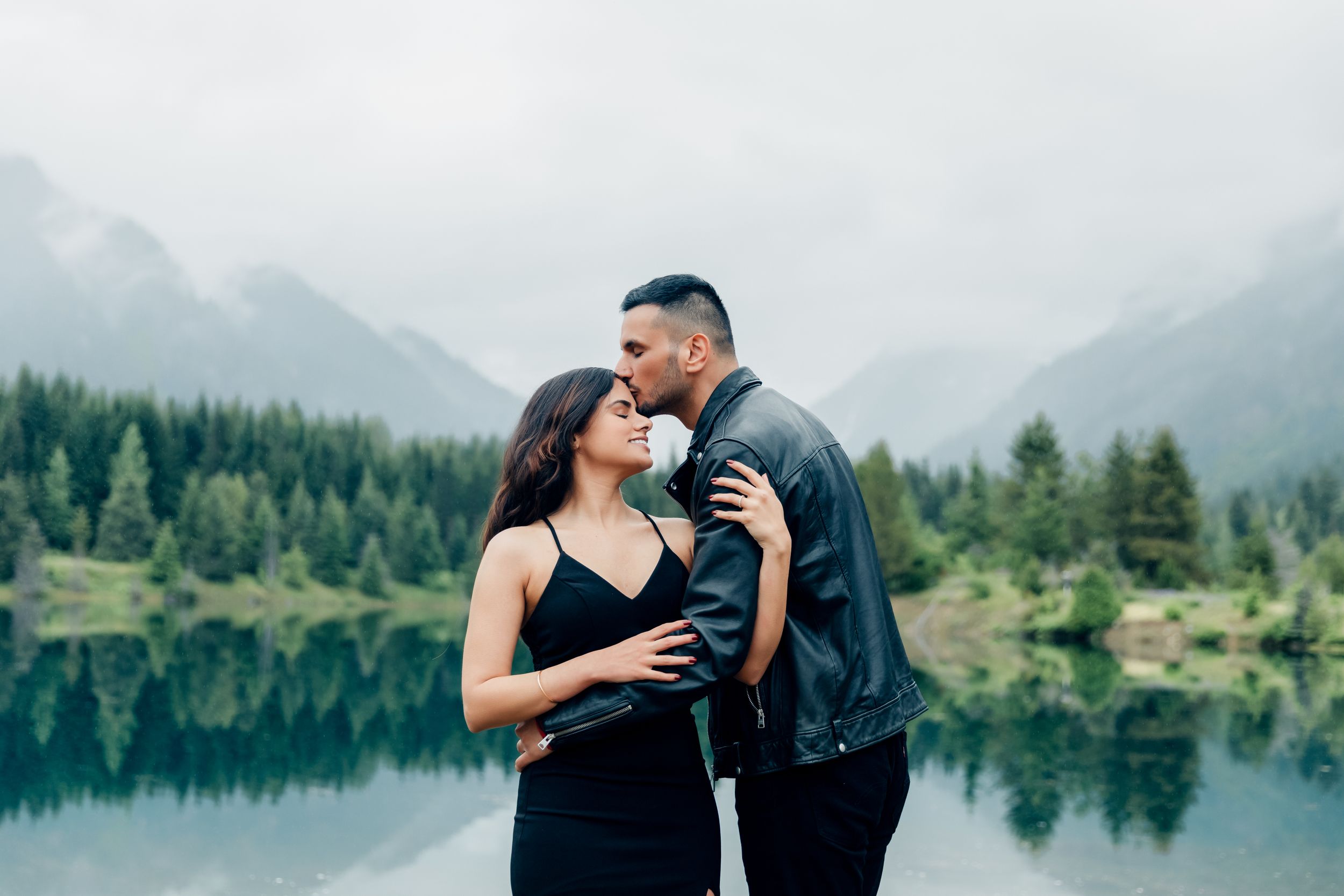 A romantic couple embraces by a serene mountain lake surrounded by evergreen forest and misty peaks.