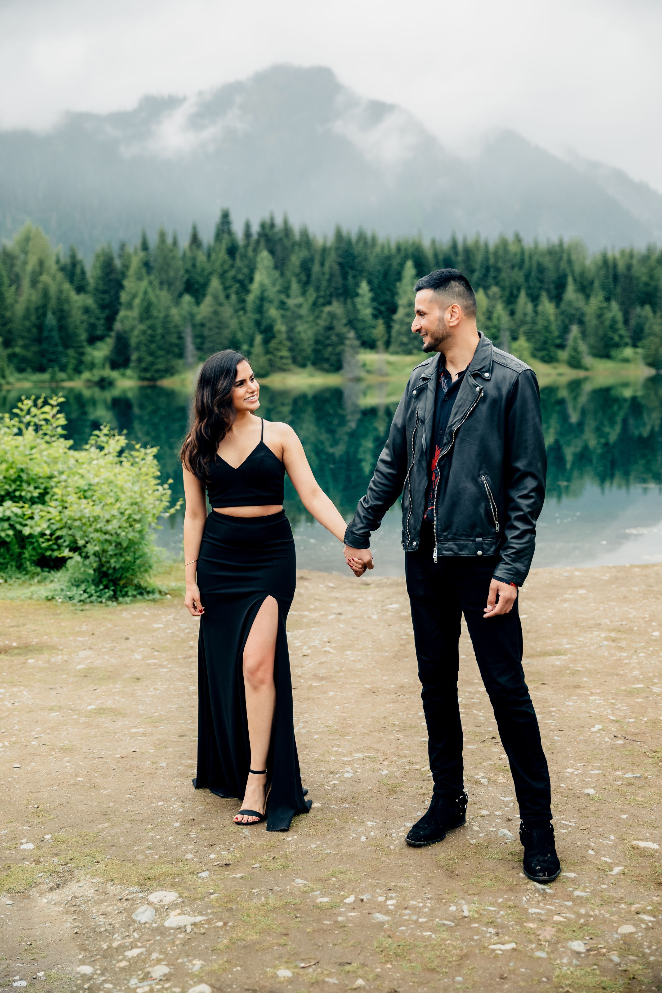 A couple in black outfits poses together on a lakeside trail with mountains and forest in the background.