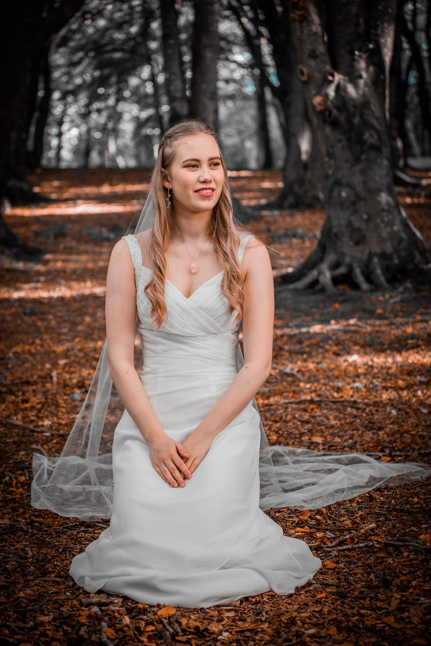 Beautiful bride in a white wedding gown kneeling gracefully among autumn leaves in a forest.