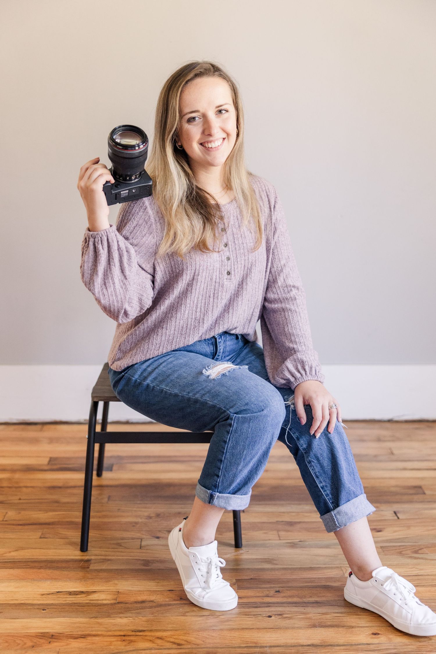 Professional photographer sits casually on chair holding camera wearing purple sweater and ripped jeans.