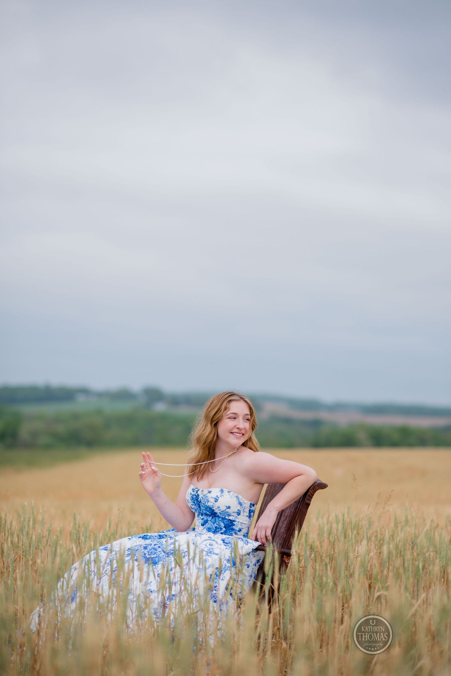 Woman in a flowing blue dress poses gracefully in an armchair among wheat fields.