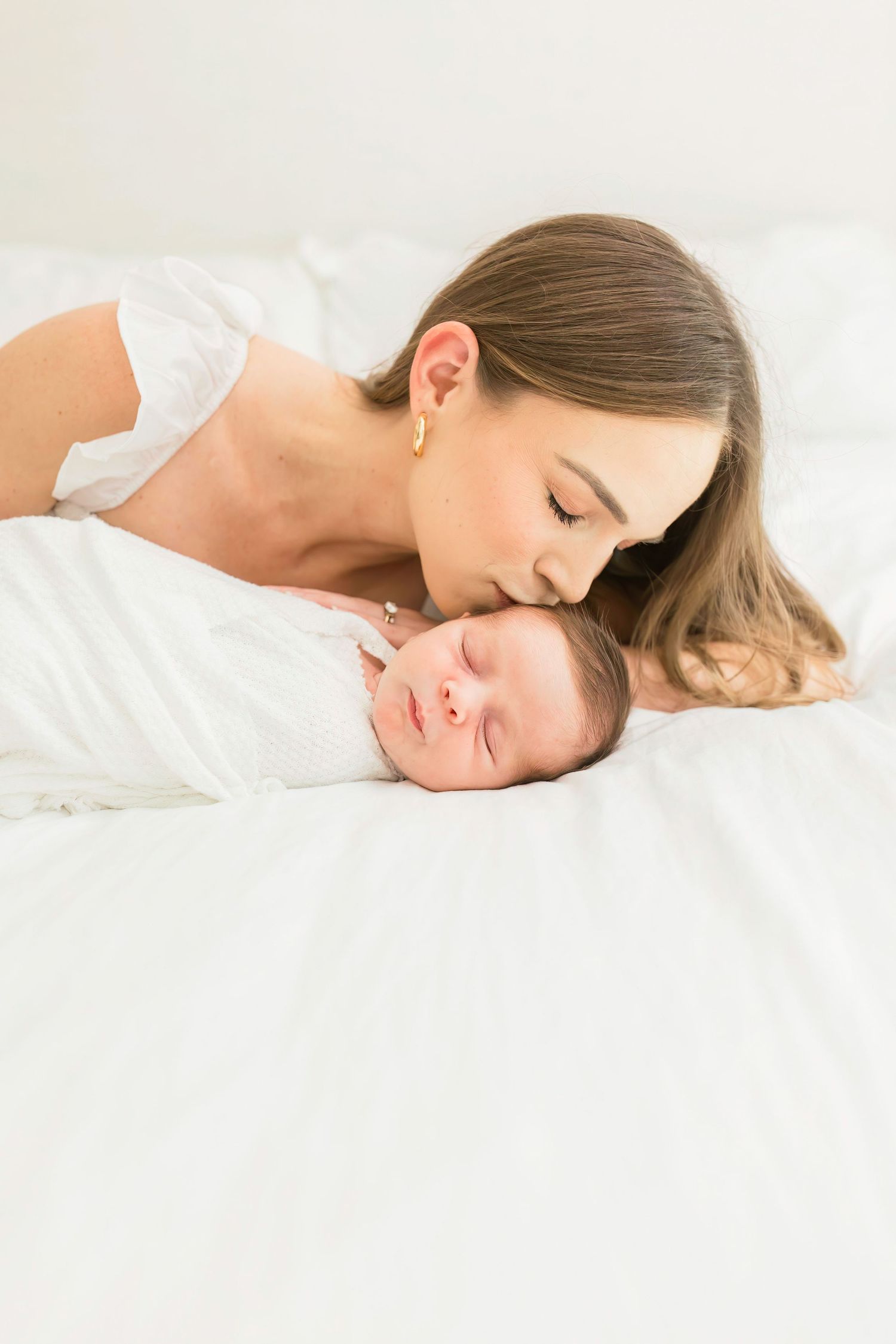 Parent sharing tender moment with sleeping newborn on white bed.