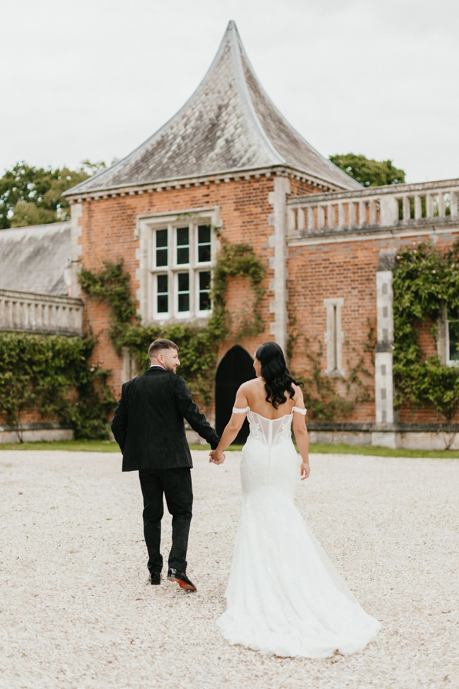 A couple walking hand in hand on a gravel path towards a historic brick building with ivy-covered walls.