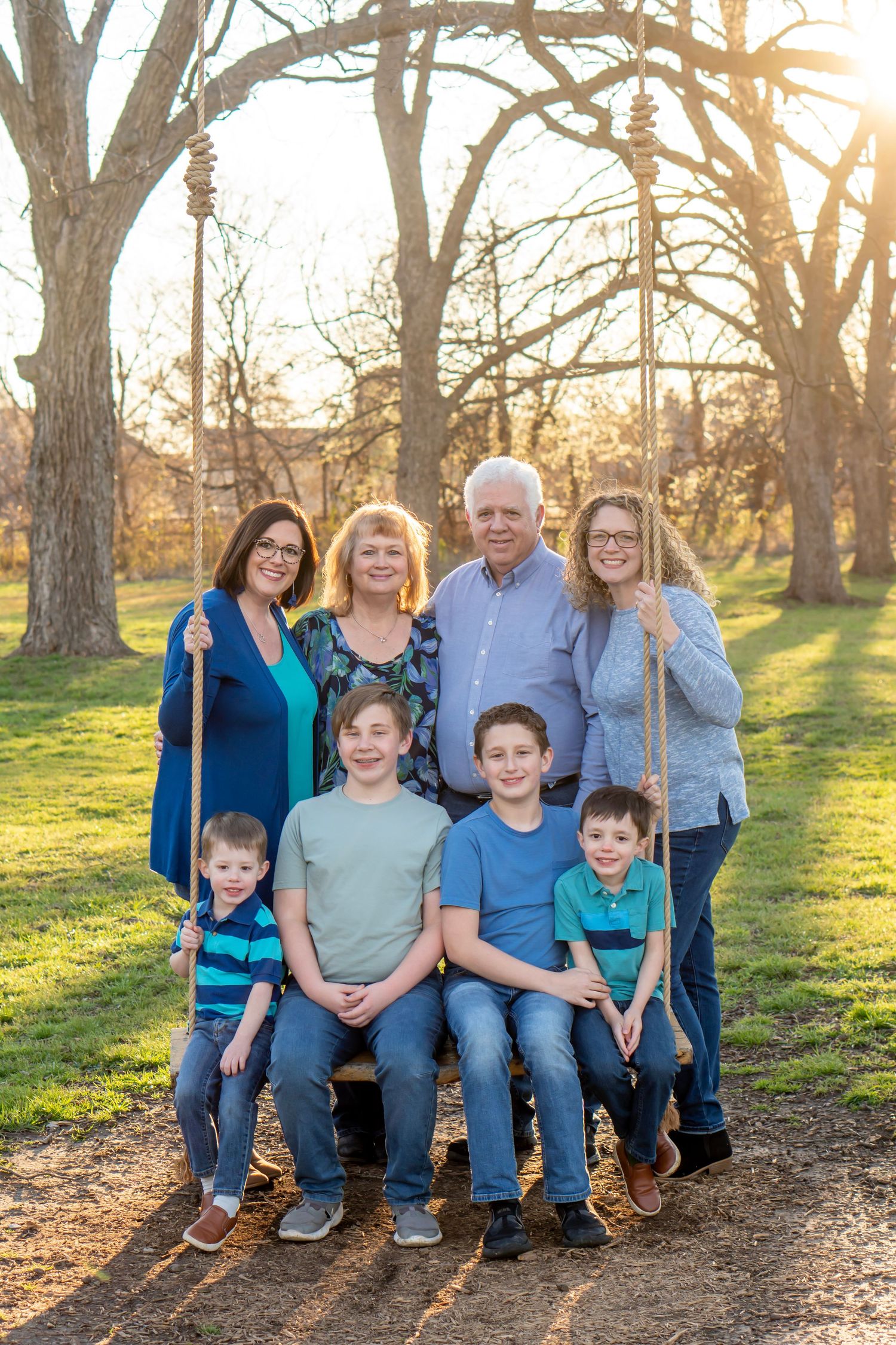 Multi-generational family portrait on a wooden swing at golden hour with bare trees in background.