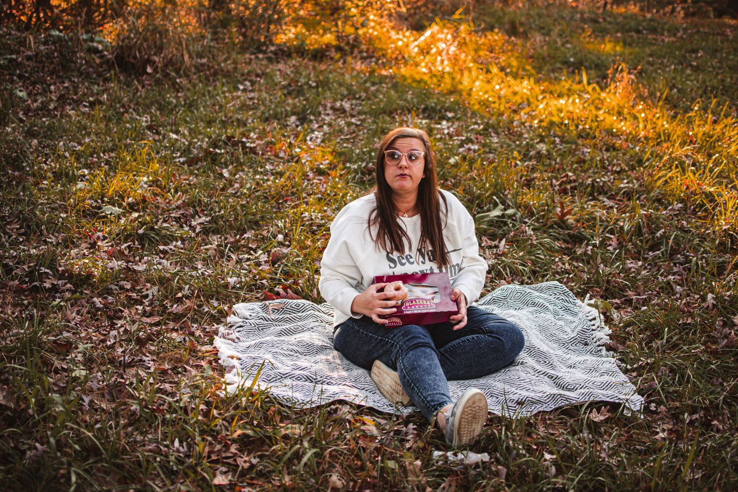 Person holds beverage while sitting comfortably on blanket in autumn woods.