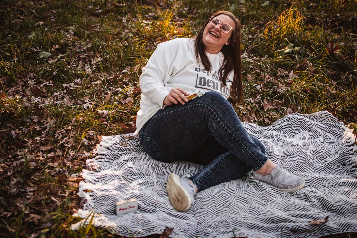 Person reclines on gray blanket surrounded by fallen leaves in autumn forest setting.