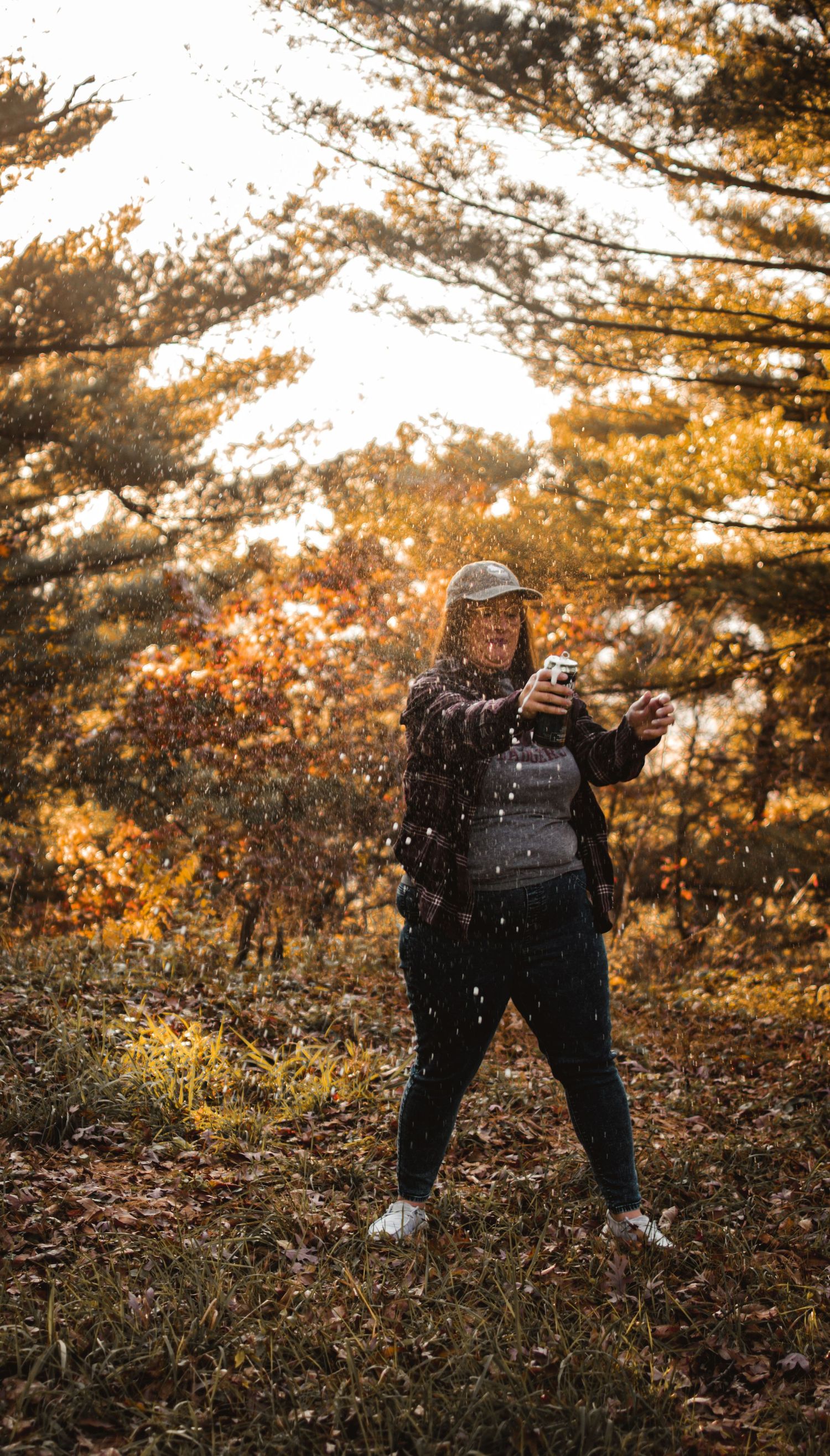 Person wearing black outfit poses in autumn forest with golden sunlight filtering through trees.